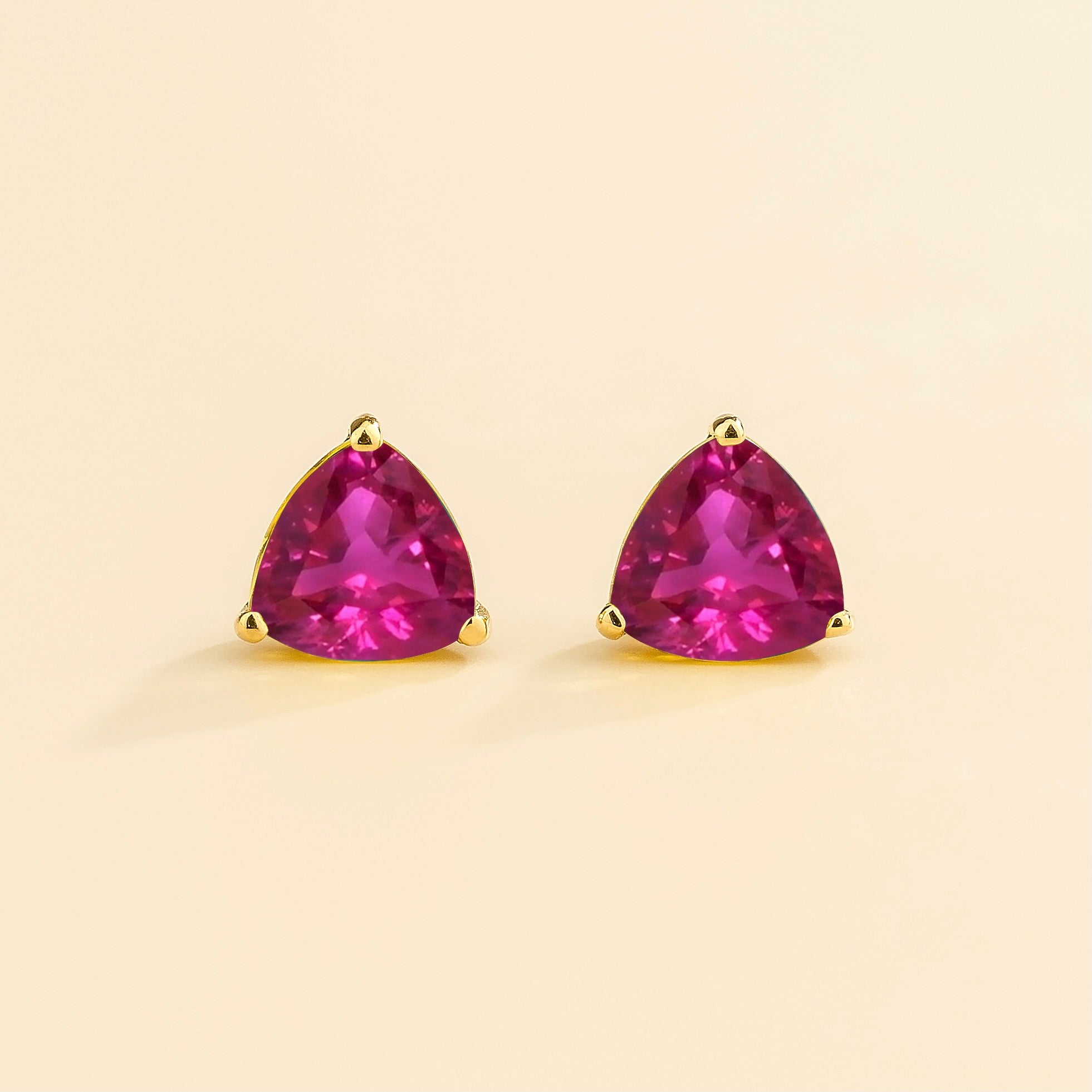 A pair of vivid pink sapphire trillion gold earrings set in yellow gold frames on a plain background.