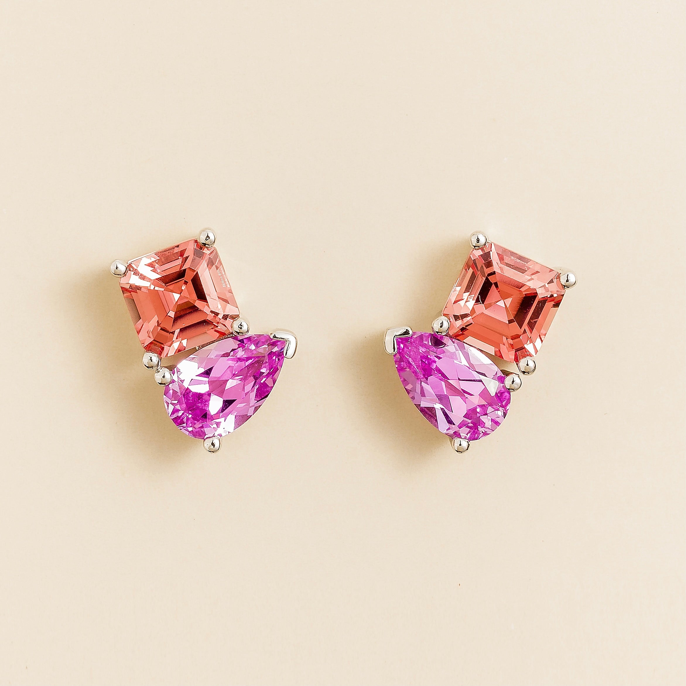 Pair of white gold earrings with pear shape pink and asscher shape padparadscha sapphire gemstones on a beige background