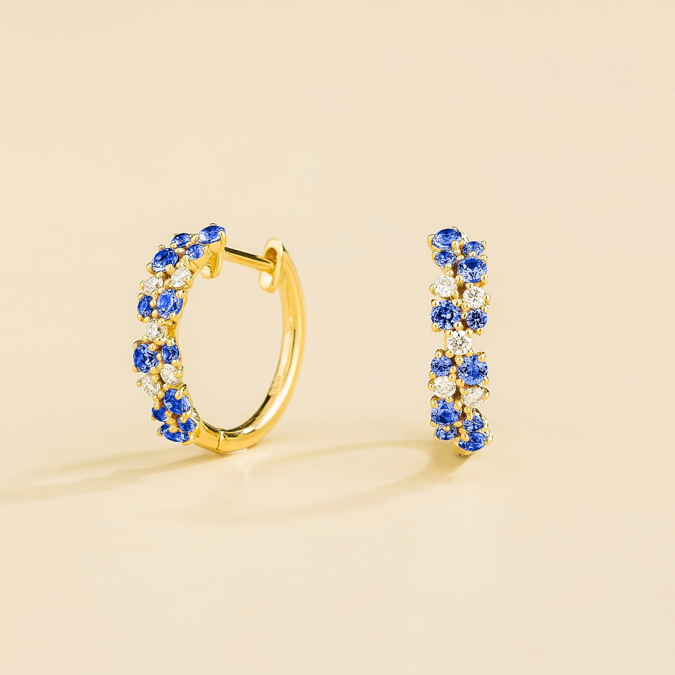 A pair of gold huggie earrings set with cluster of round blue sapphire and diamond accents, displayed against a neutral background.