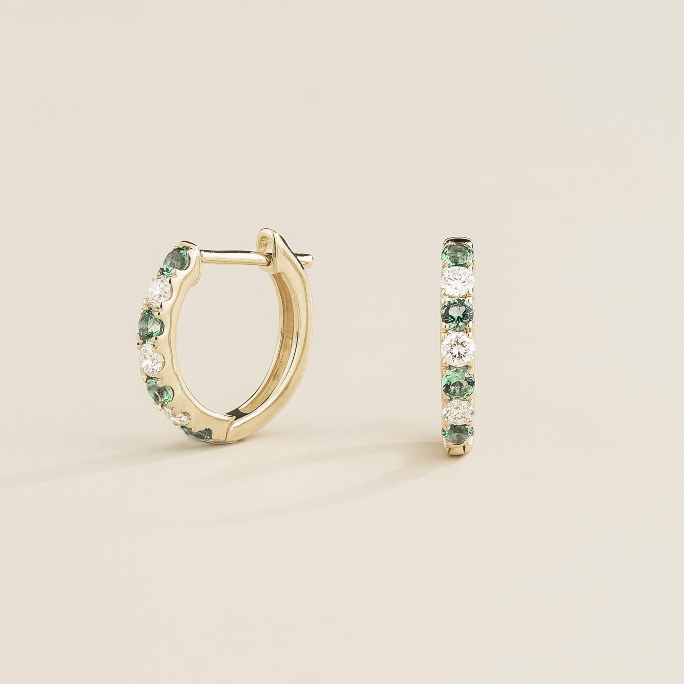 A pair of white gold huggie earrings set with emeralds and diamonds, displayed against a neutral background.