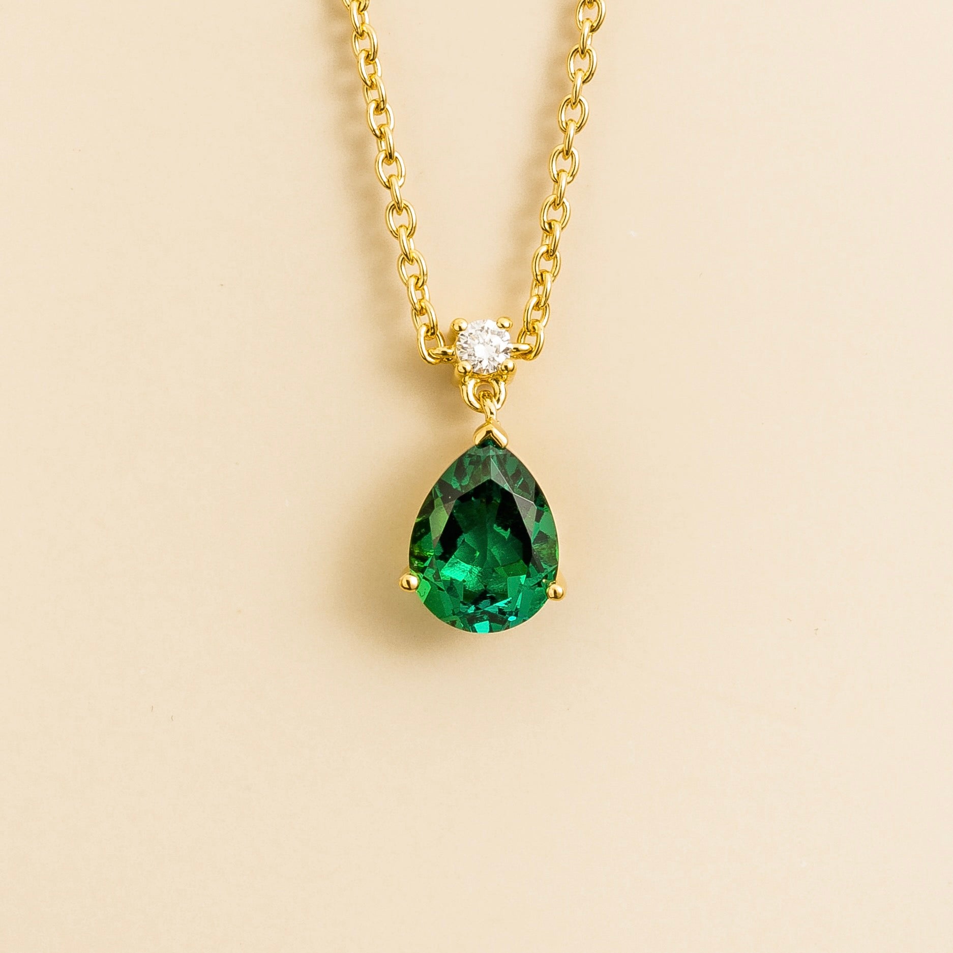 A classic pendant necklace with a pear shaped emerald and a smaller round diamond set in gold, displayed against a beige background.