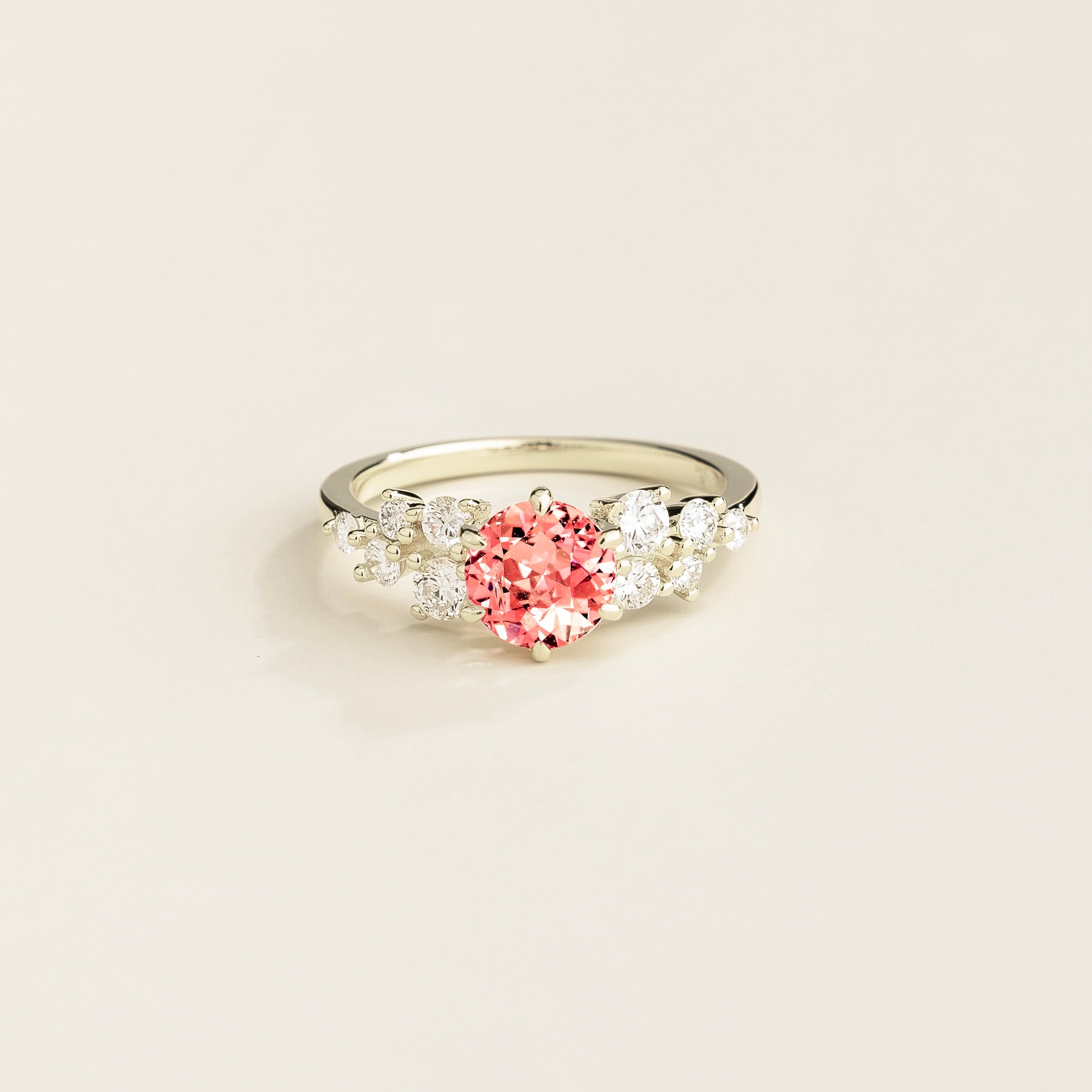 A white gold ring with a padparadscha sapphire center stone and smaller diamonds cluster set in gold, displayed against a neutral background.