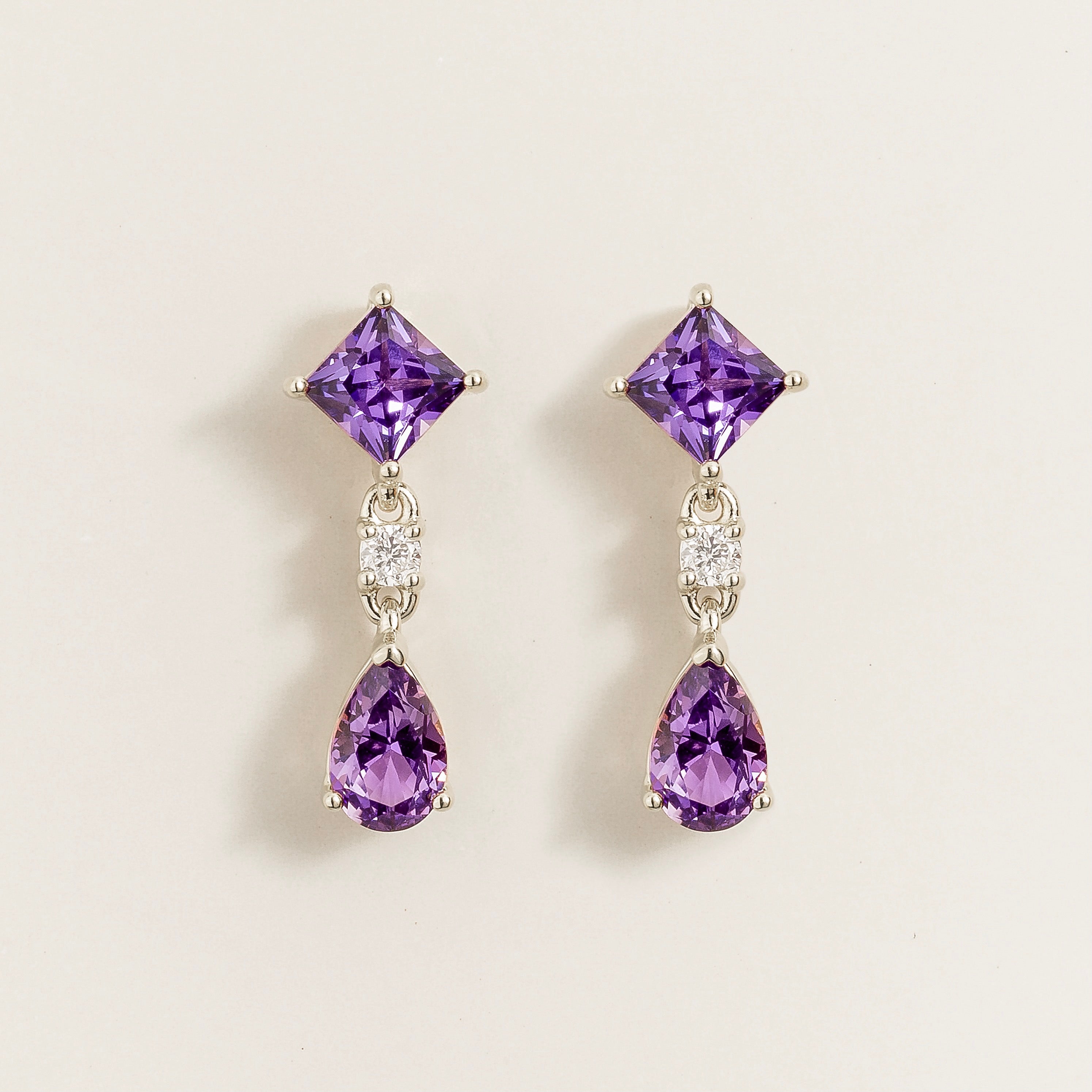 A pair of white gold earrings featuring princess cut purple sapphire atop a pear cut purple sapphire, with smaller round cut diamonds in between.