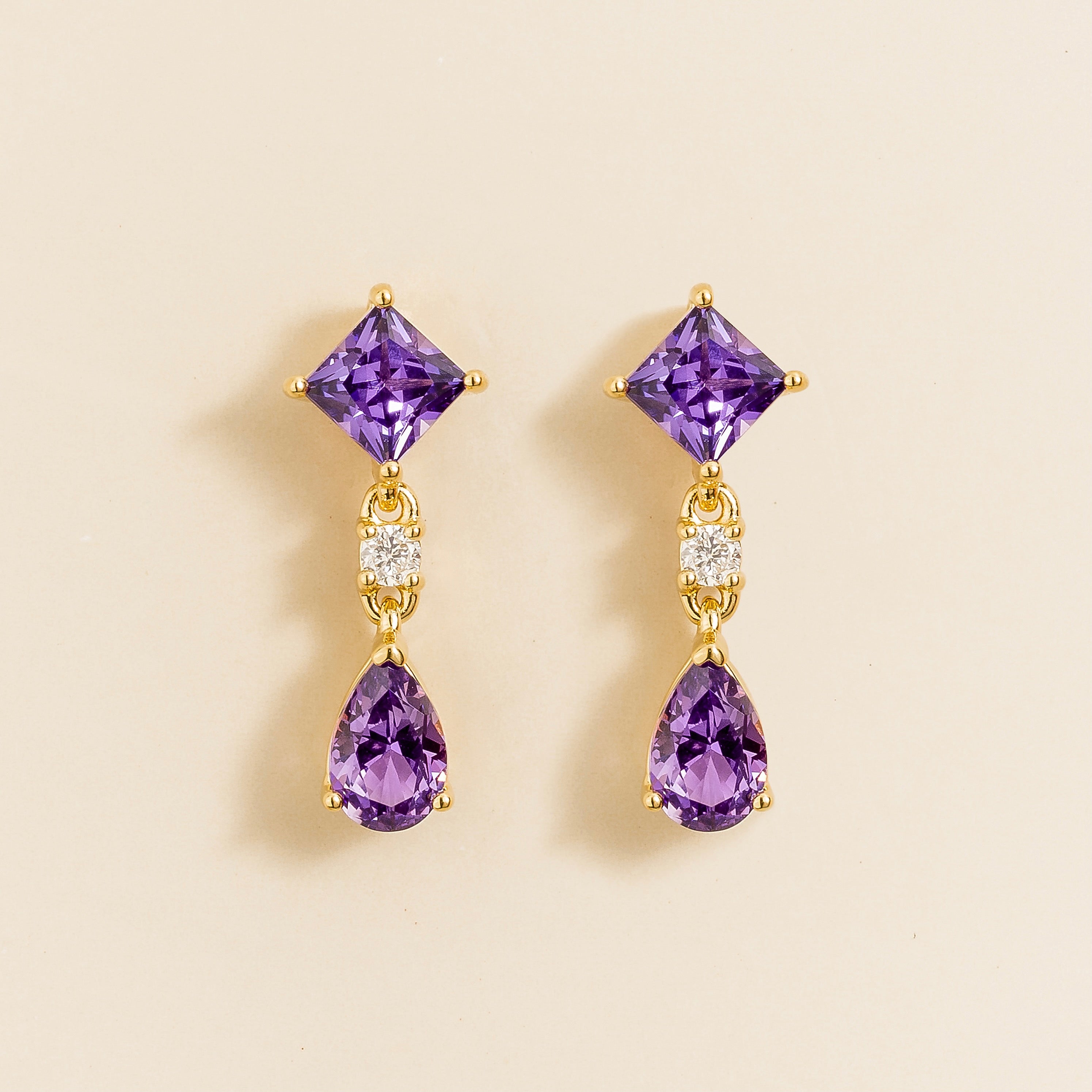 A pair of yellow gold earrings featuring princess cut purple sapphire atop a pear cut purple sapphire, with smaller round cut diamonds accenting the design.