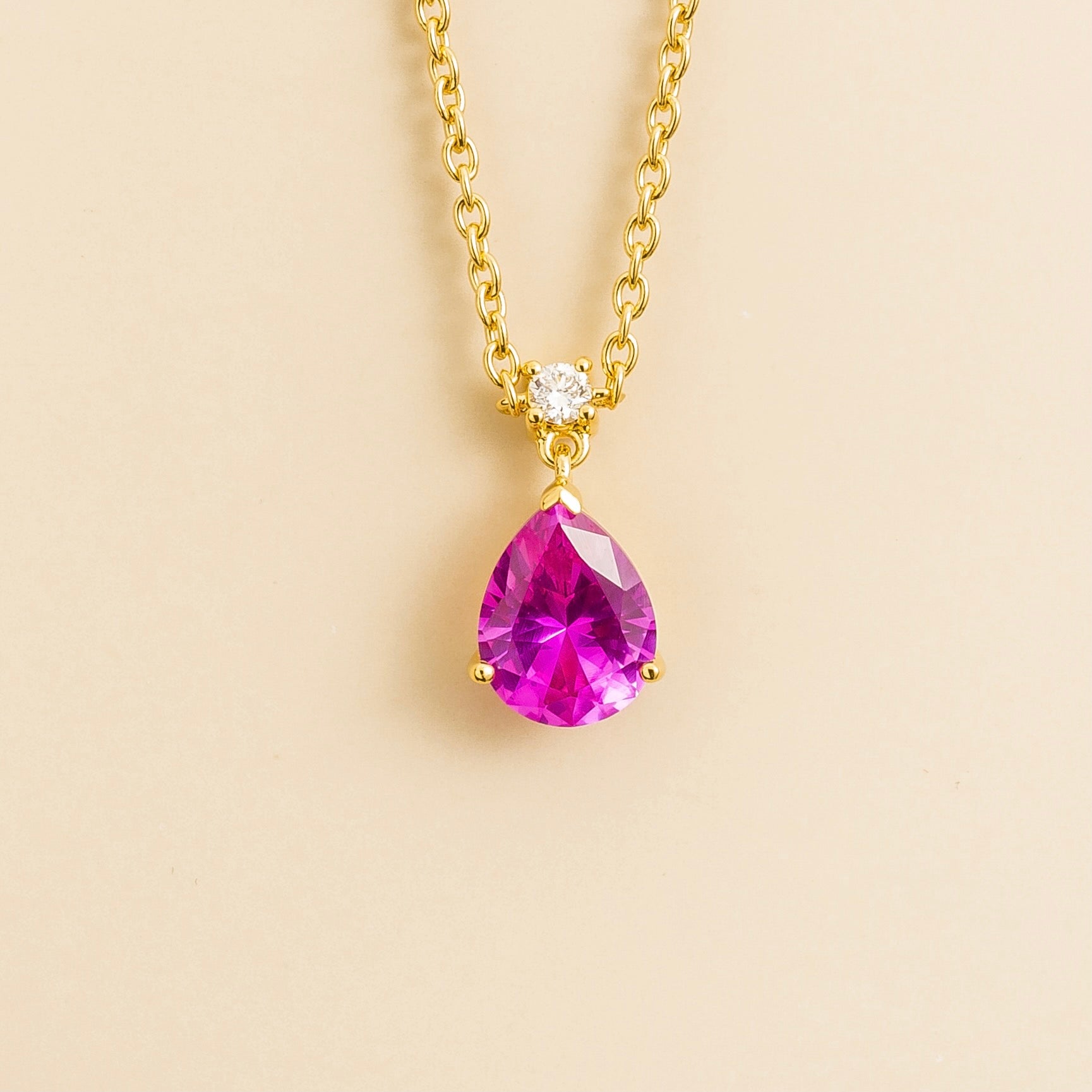 A classic gold necklace with a pear shape vivid pink sapphire pendant and a smaller diamond, displayed against a neutral background.