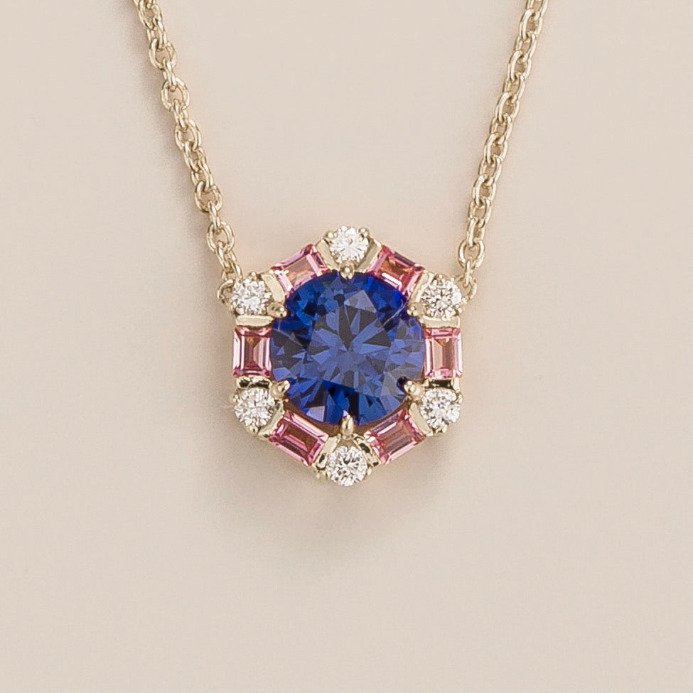 A white gold necklace set with a round blue sapphire, pink sapphire, and diamond halo pendant in a hexagon shape.
