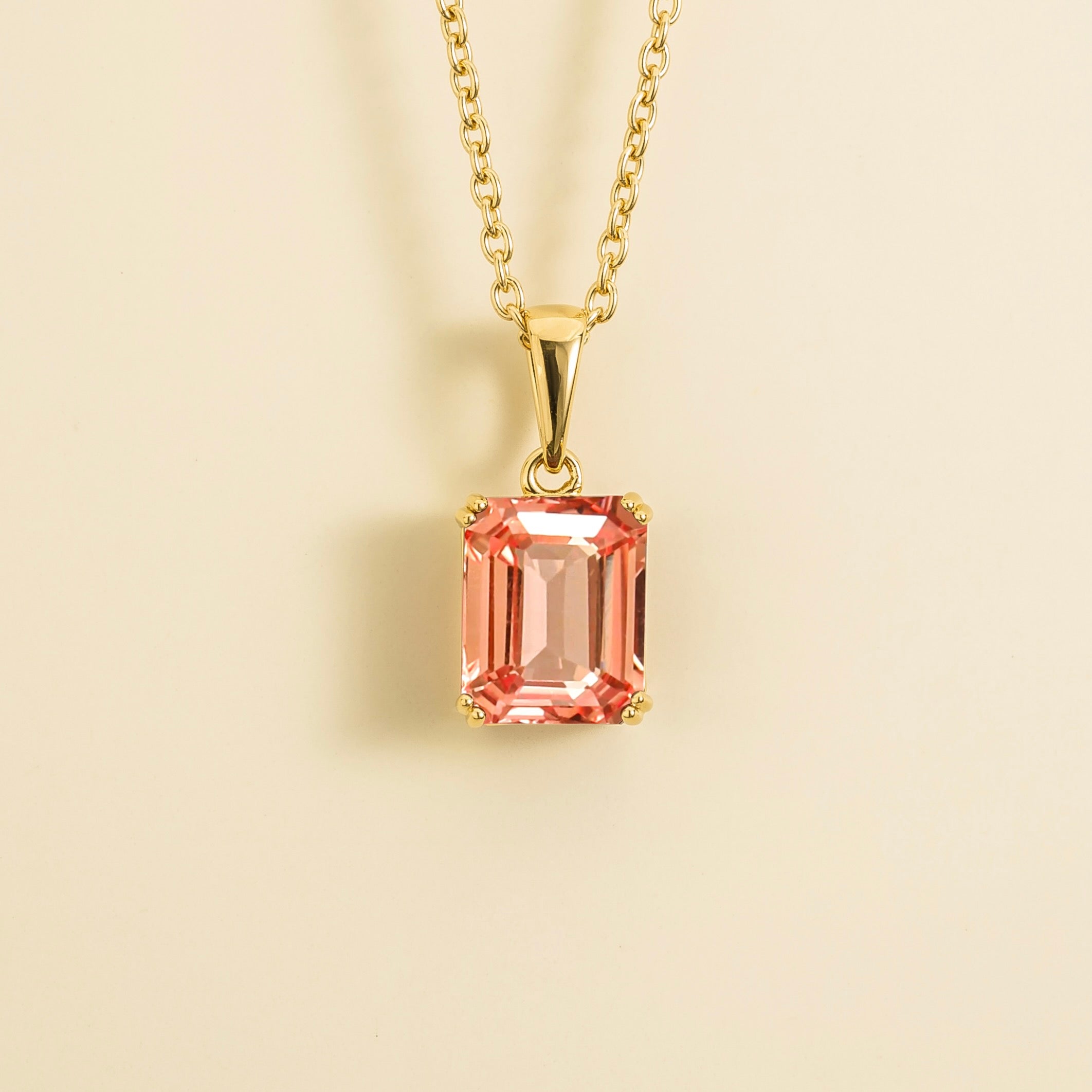 A gold pendant necklace with an emerald-cut Padparadscha sapphire centerpiece, displayed against a light background.