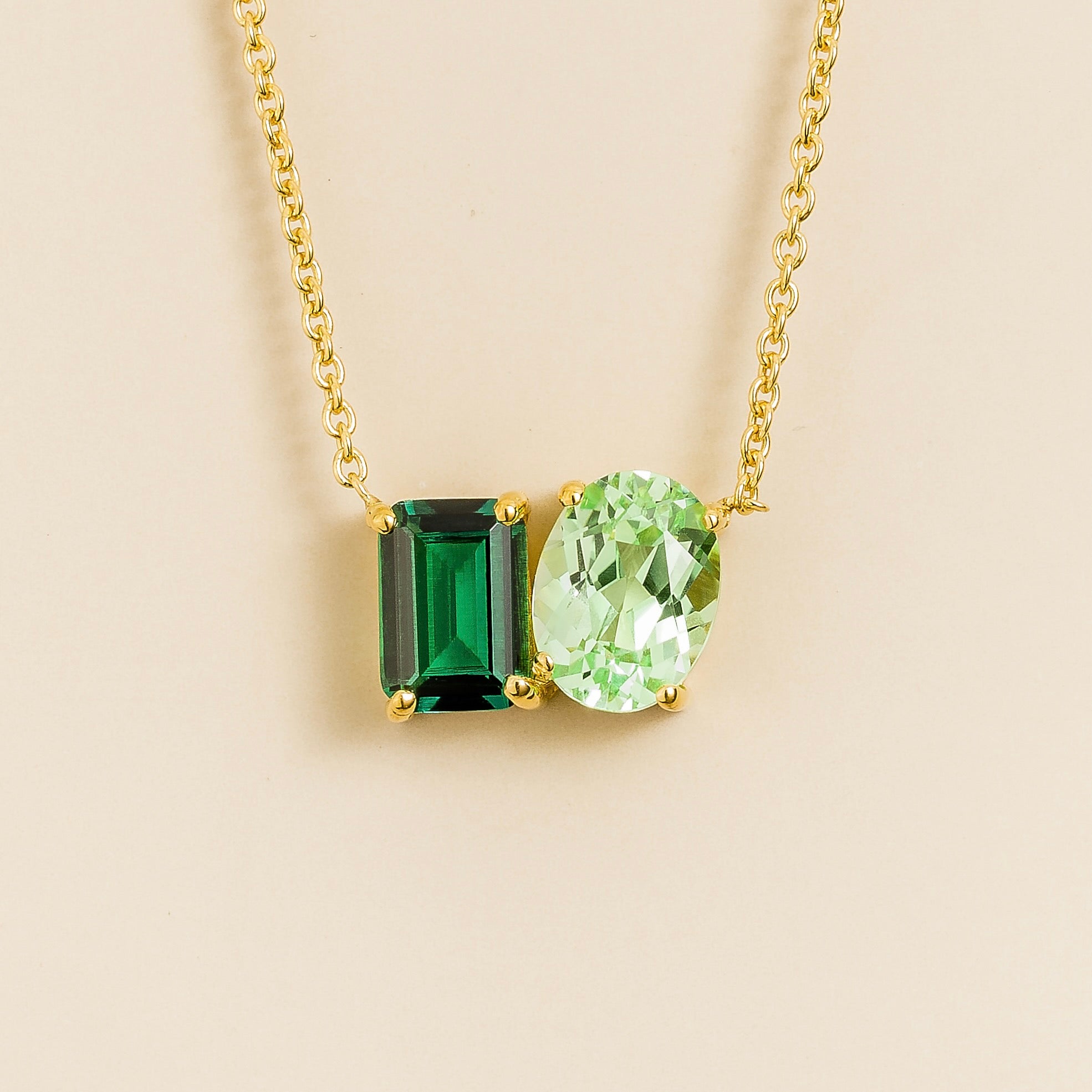 Gold Toi et Moi necklace with an emerald cut green emerald and an oval green sapphire pendant on a light background.