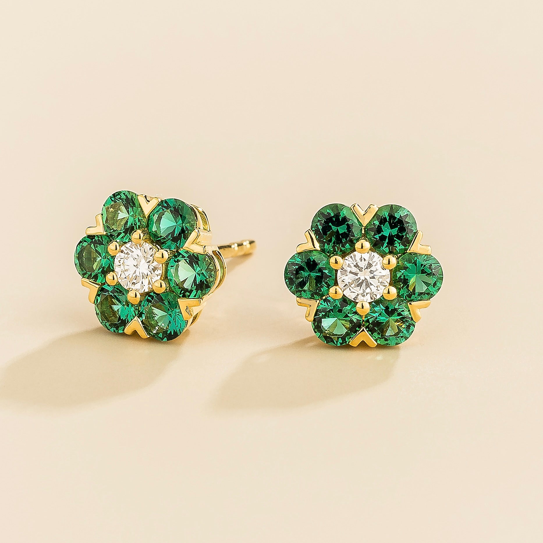 A pair of gold earrings featuring a central diamond with emerald accents arranged in a floral daisy pattern.