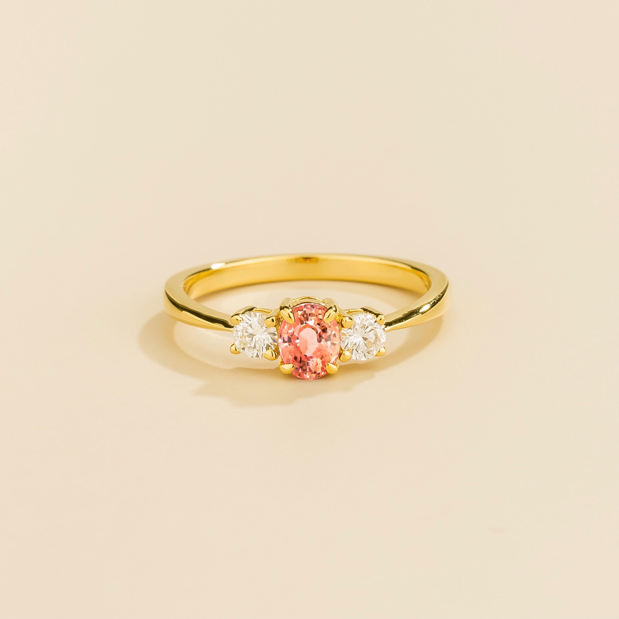 A gold ring featuring a central Padparadscha sapphire flanked by two diamonds, set against a neutral background.
