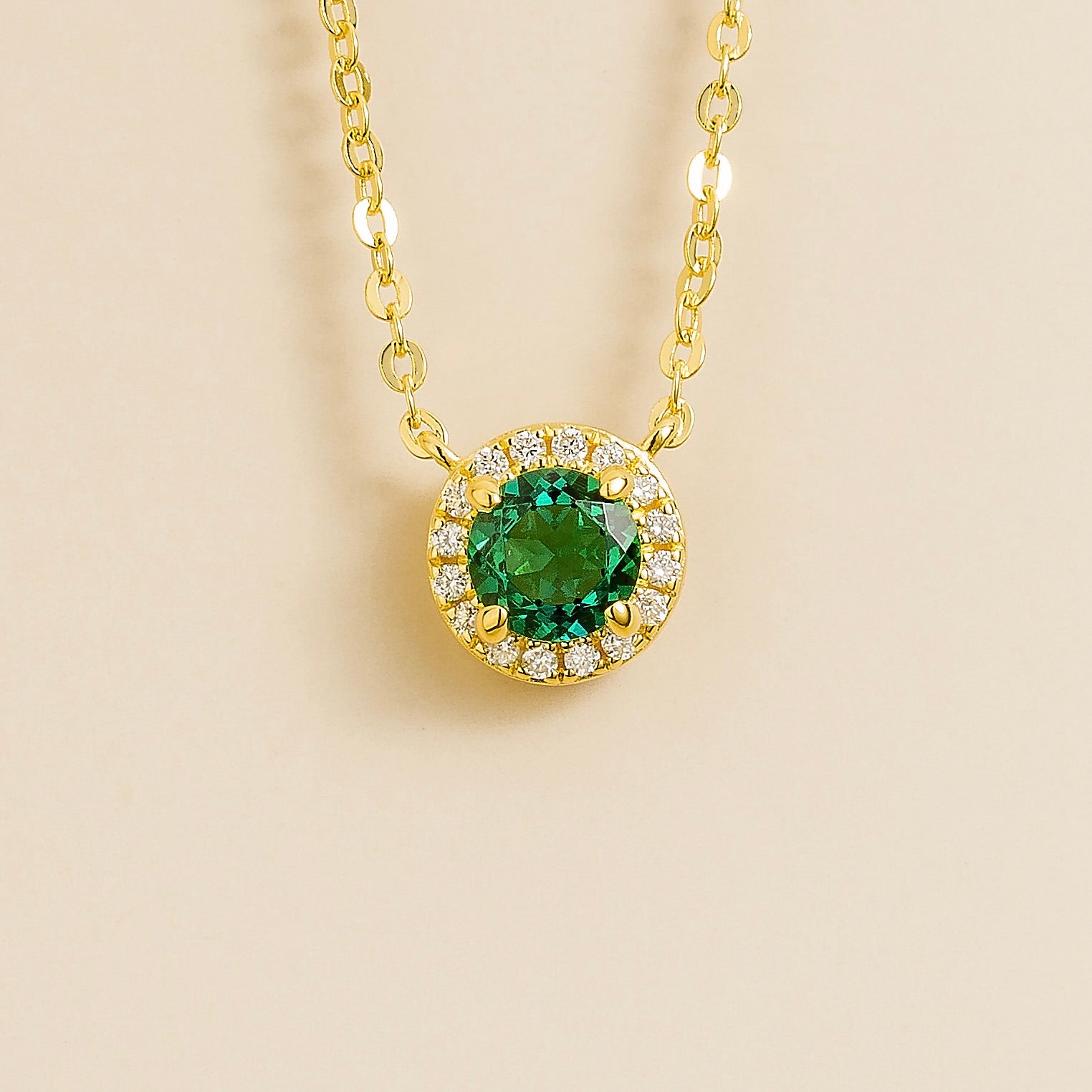 A gold necklace with a halo design featuring a round emerald centerpiece surrounded by smaller diamonds, displayed against a light background.