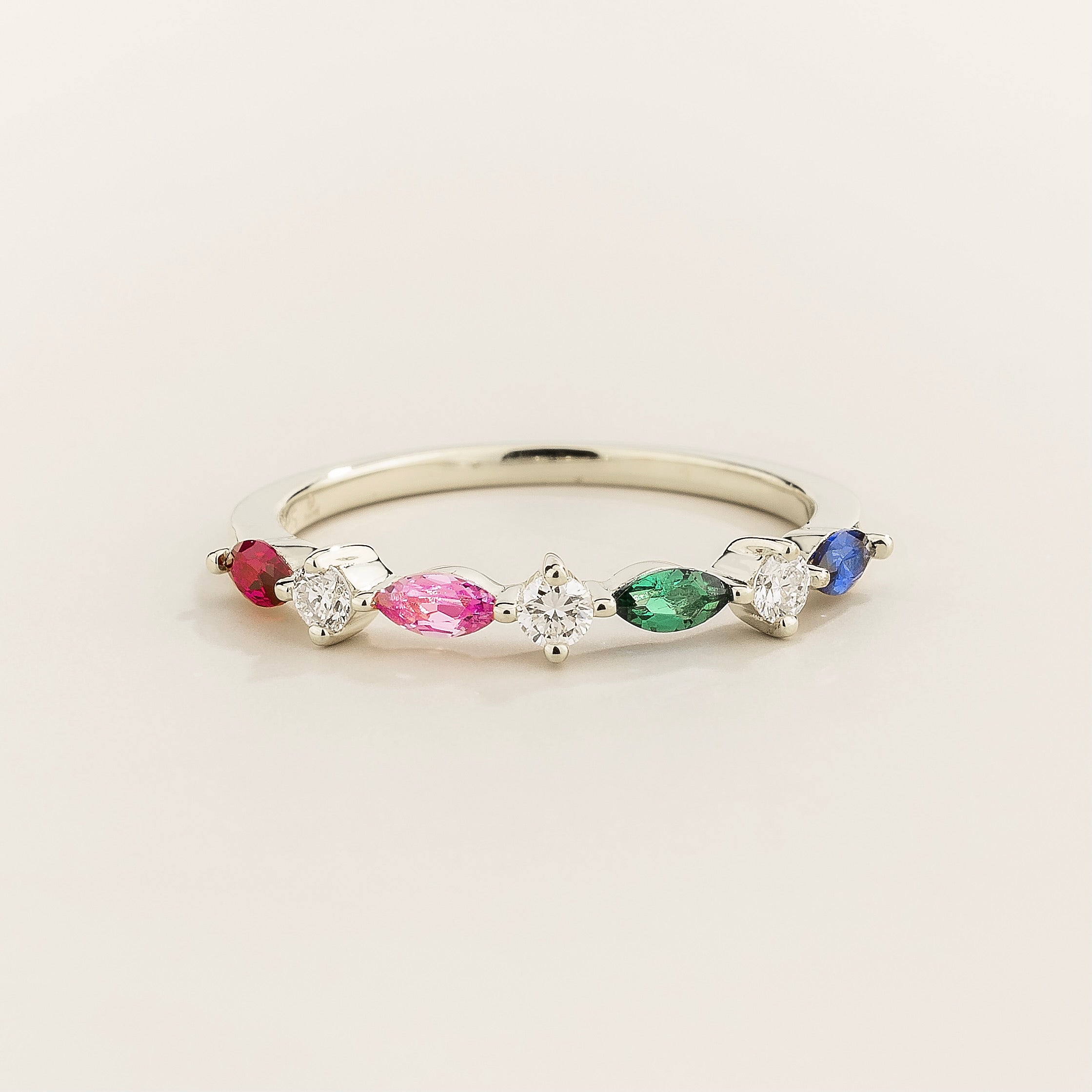 A white gold band ring with marquise cut ruby, pink sapphire, emerald, blue sapphire and round cut diamonds set in an alternating pattern on a plain