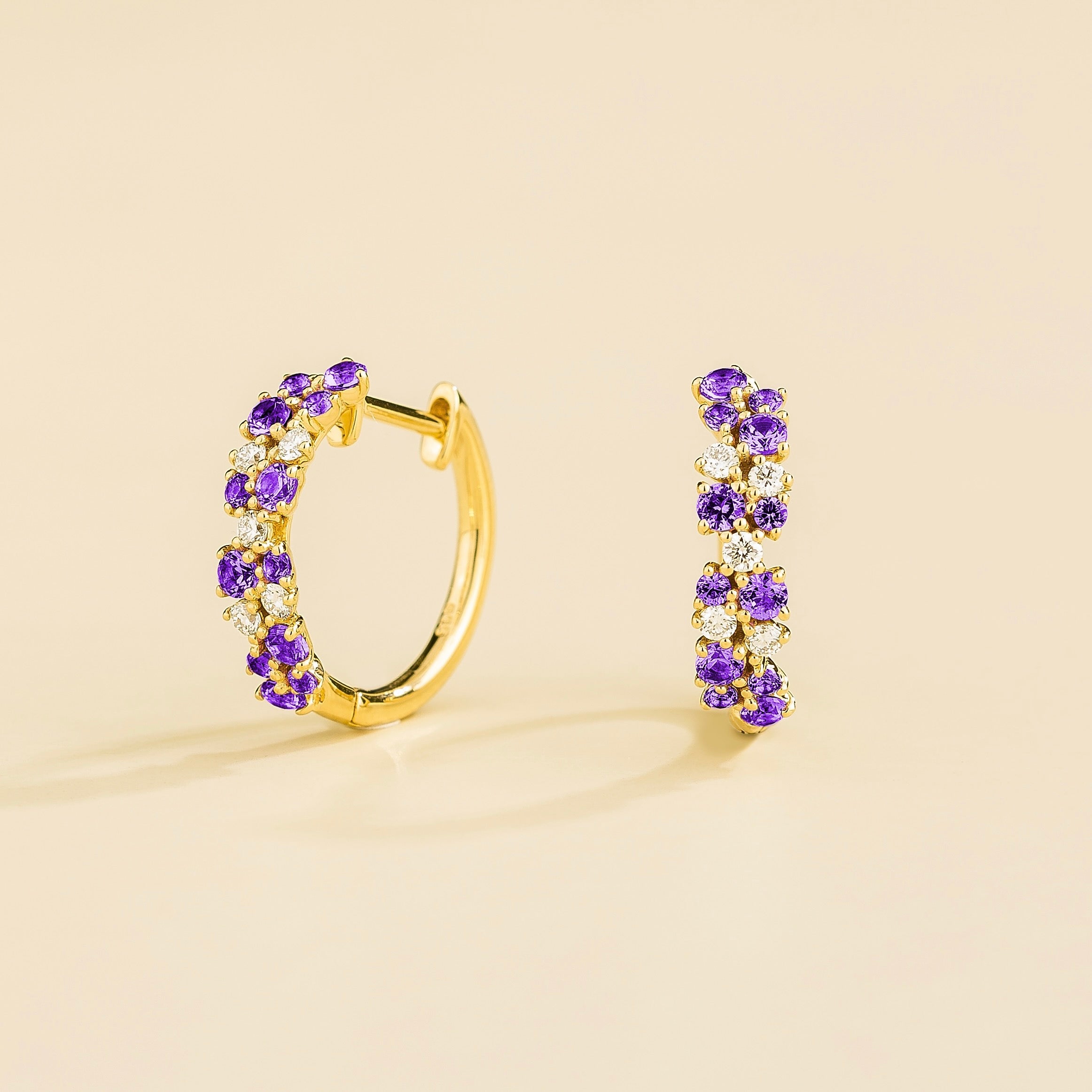 A pair of gold huggie earrings set with cluster of round purple sapphire and diamond accents, displayed against a neutral background.