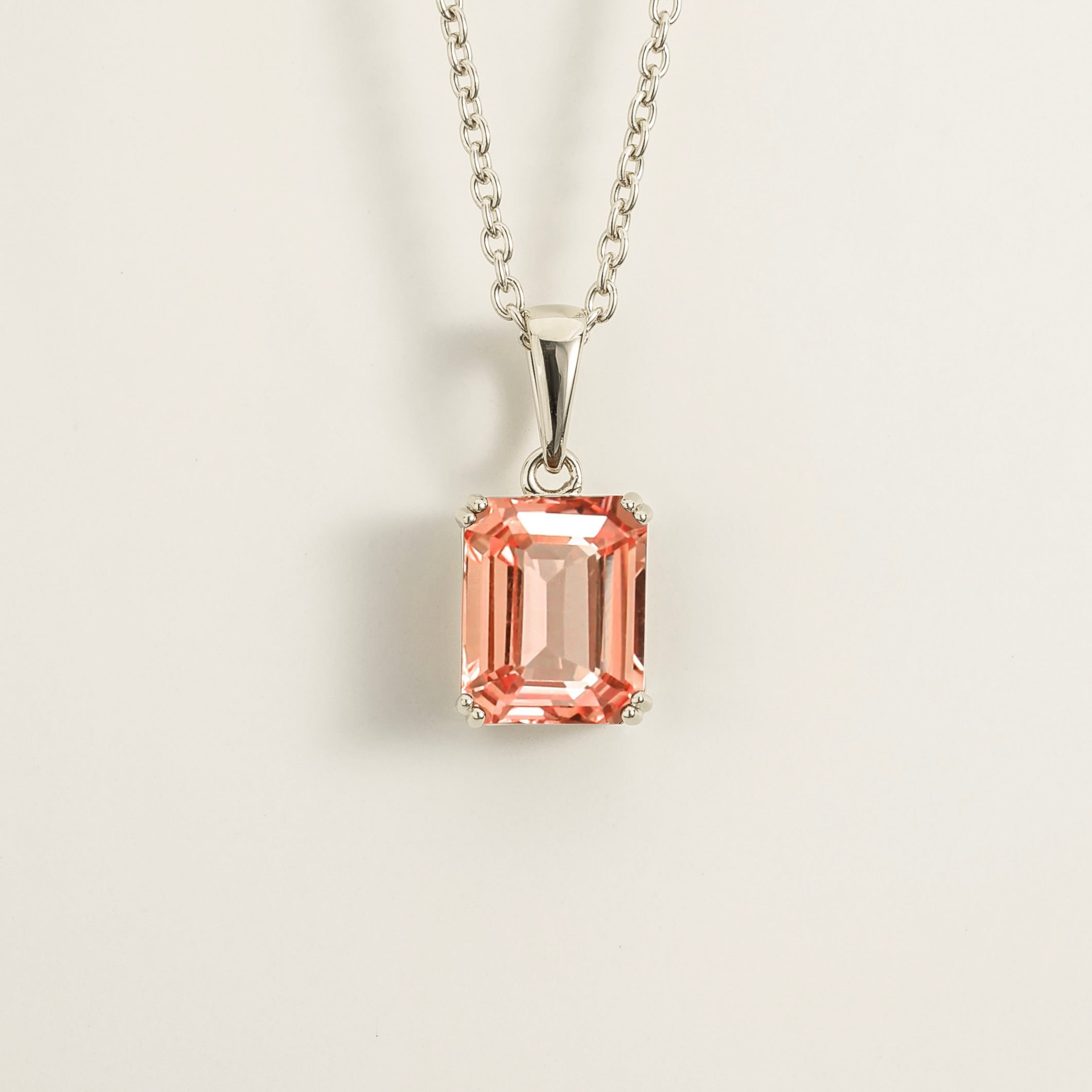 A white gold pendant necklace with a padparadscha sapphire in an emerald cut setting, attached to a white gold chain. London jewellery store