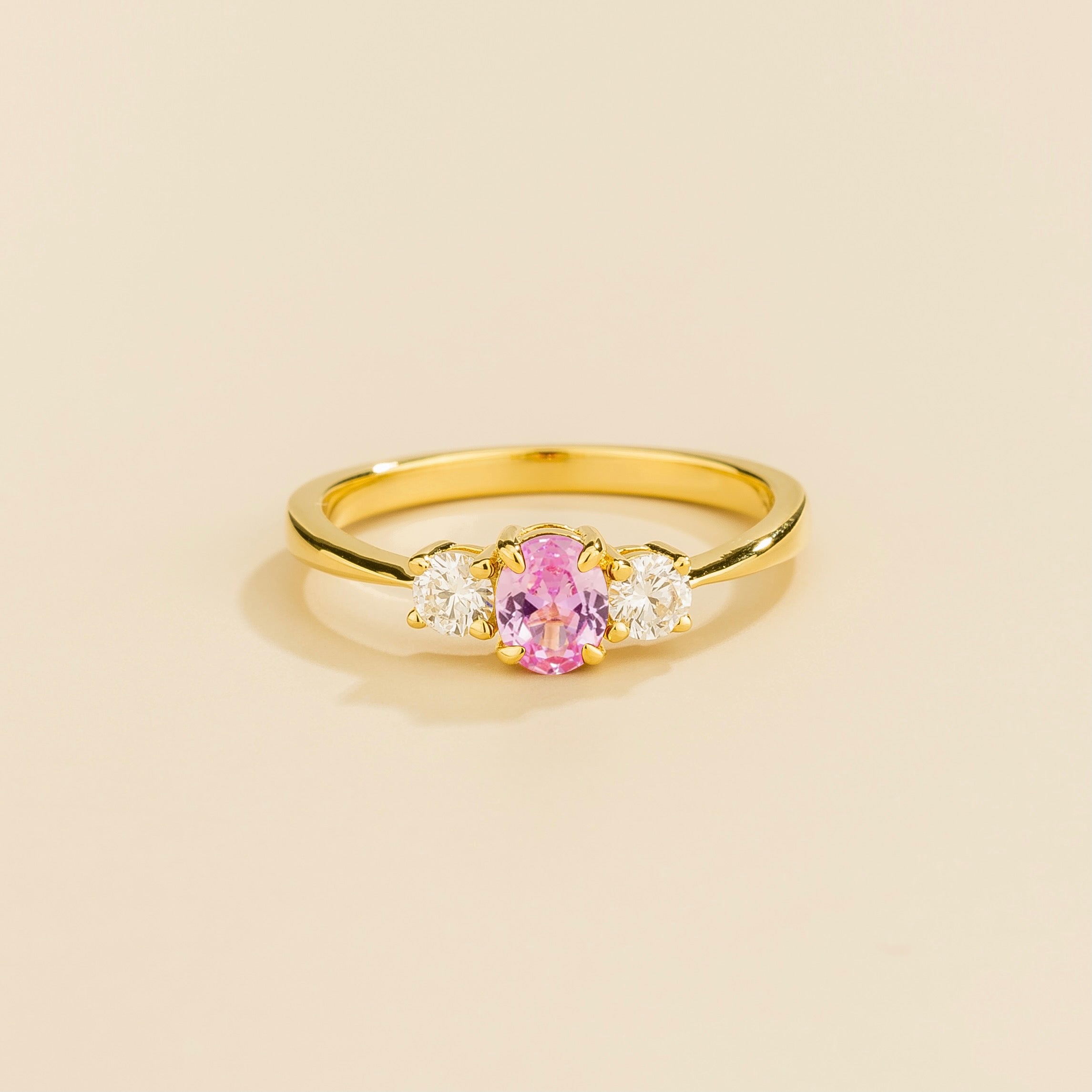 A gold ring featuring a central pink sapphire with two smaller diamonds on each side, set against a neutral background.