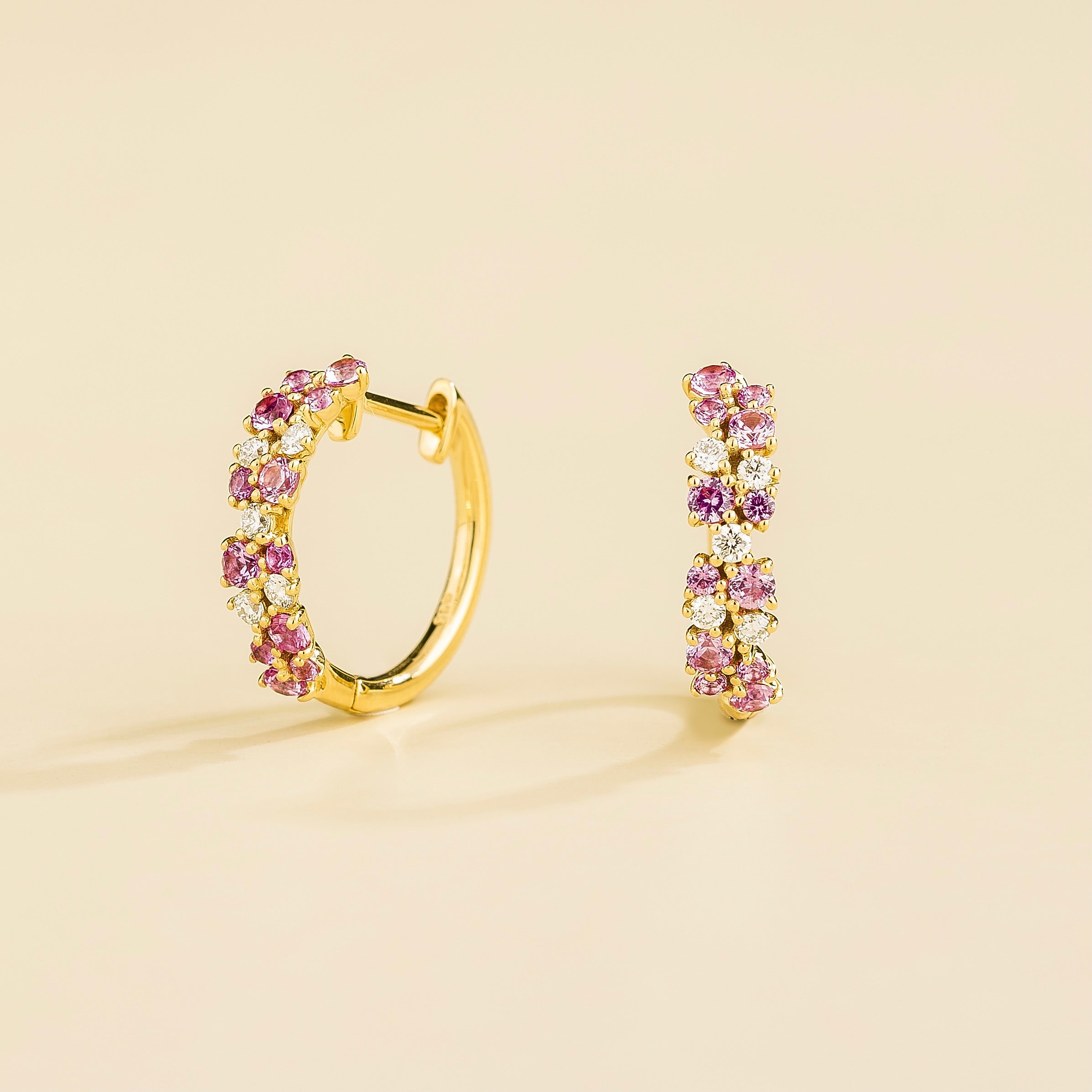 A pair of gold huggie earrings set with cluster of round pink sapphire and diamond accents, displayed against a neutral background.