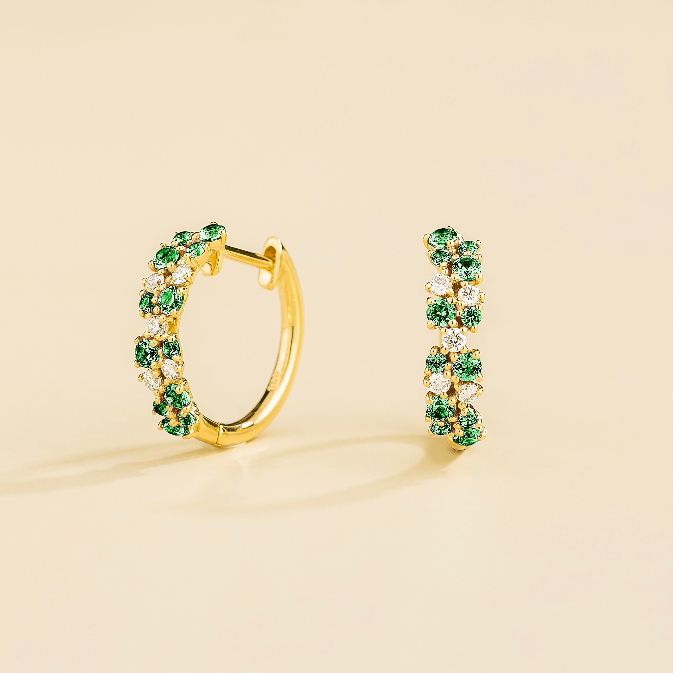 A pair of white gold huggie earrings set with cluster of round emerald and diamond accents, displayed against a neutral background.