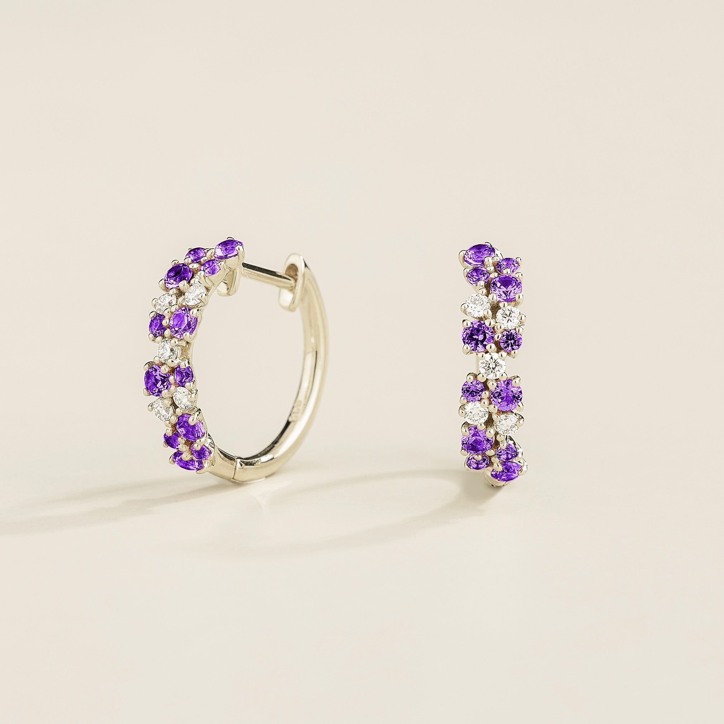 A pair of white gold huggie earrings set with cluster of round purple sapphire and diamond accents, displayed against a neutral background.