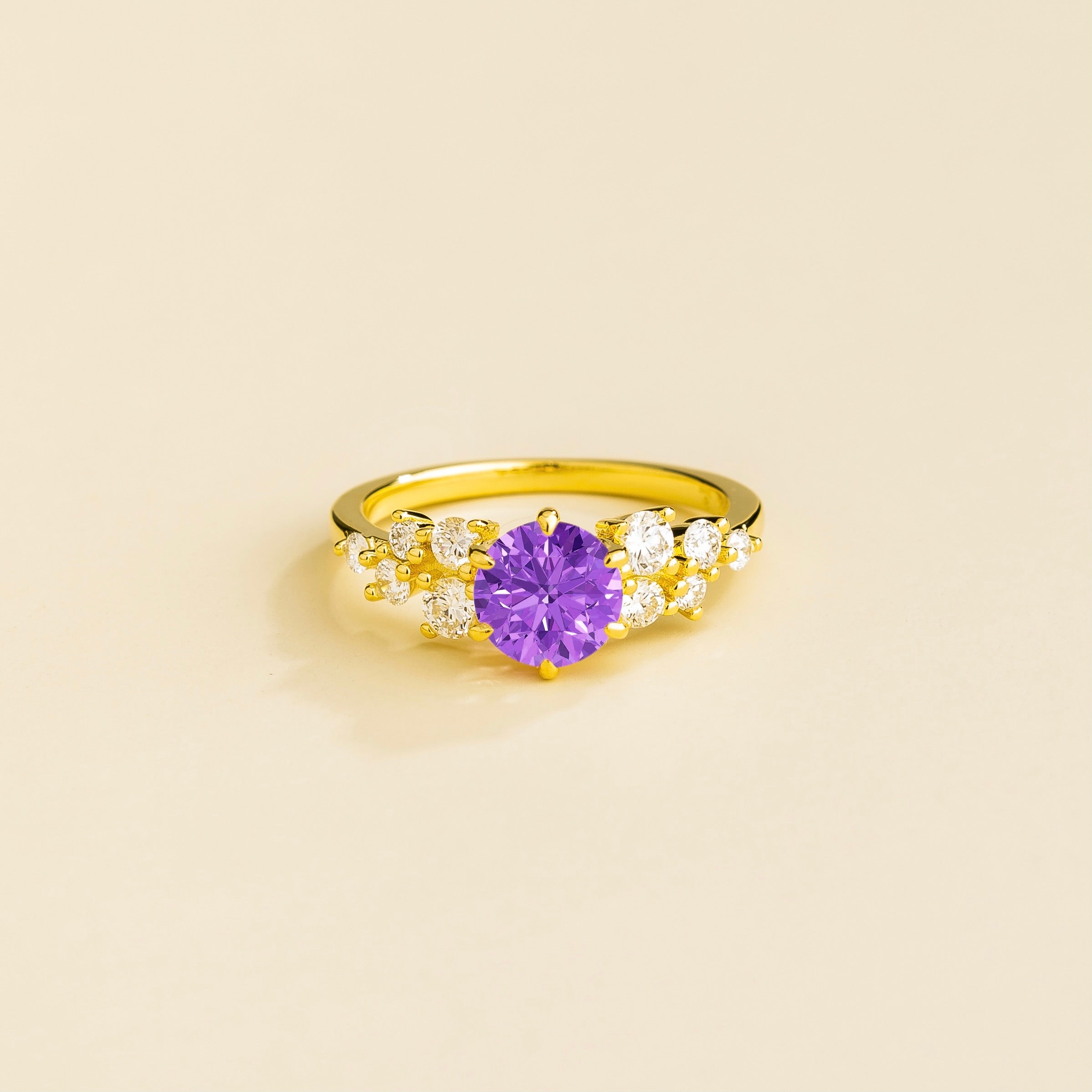 A gold ring with a purple sapphire center stone and smaller diamonds cluster set in gold, displayed against a neutral background.