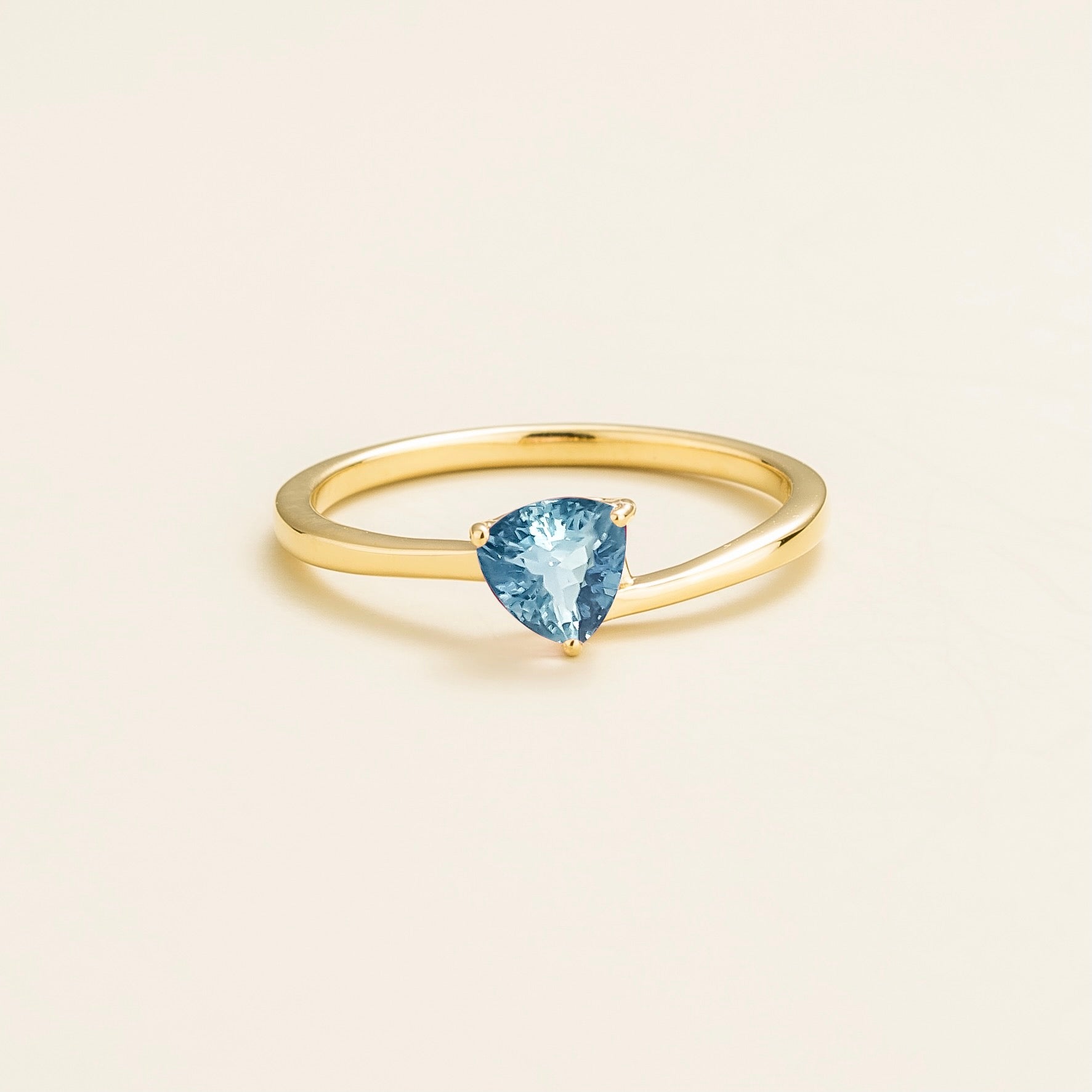 A solitaire gold ring with a trillion-cut aquamarine in a curved setting, displayed against a light background.