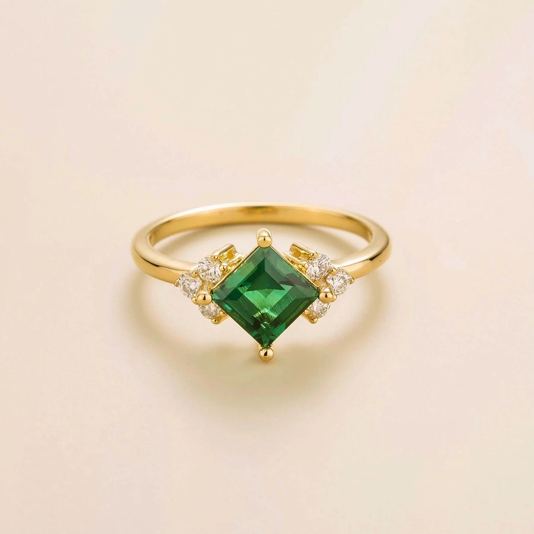 A gold ring with a central princess cut emerald and small diamond accents on a plain background.