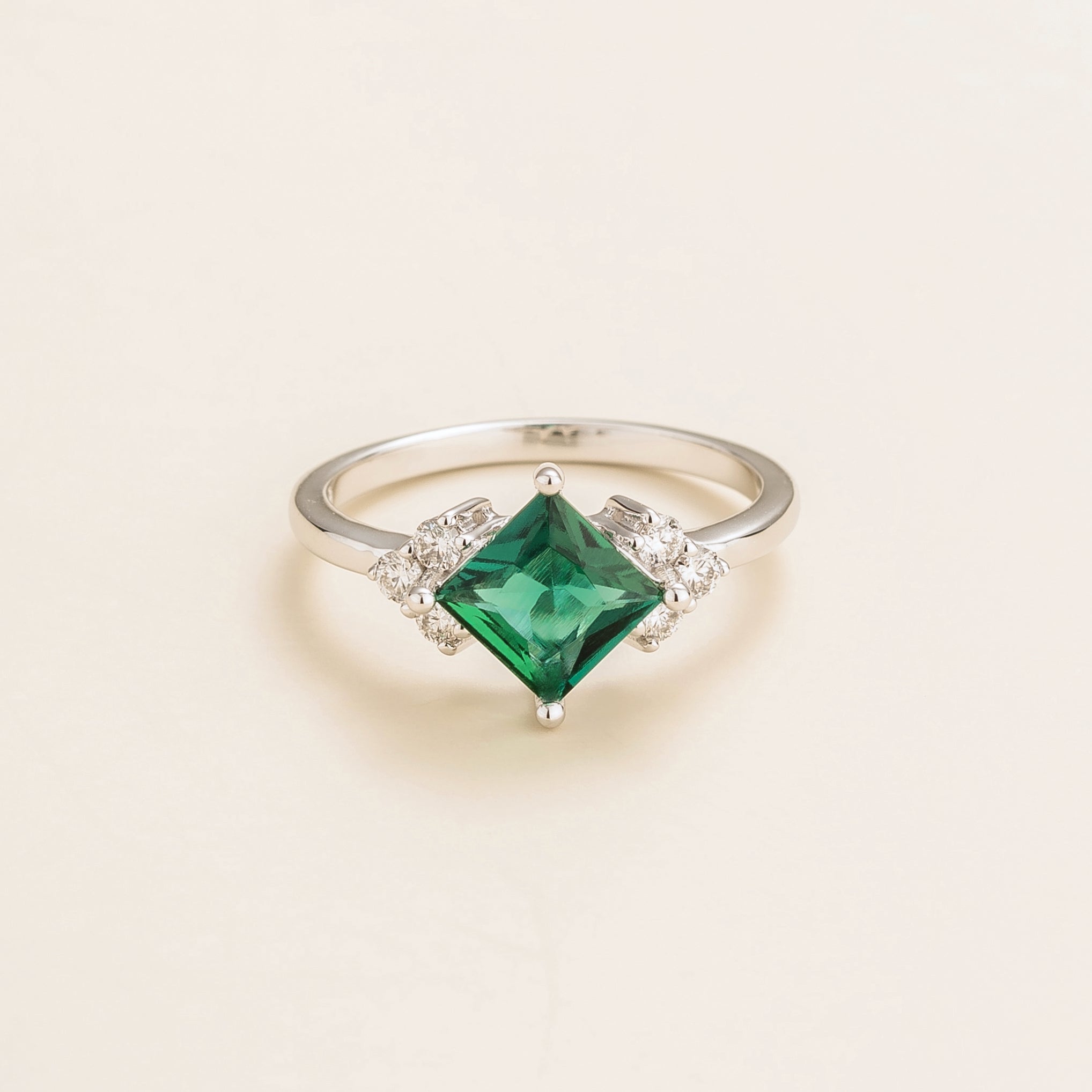 A white gold ring with a princess cut emerald center stone and smaller diamonds around it.