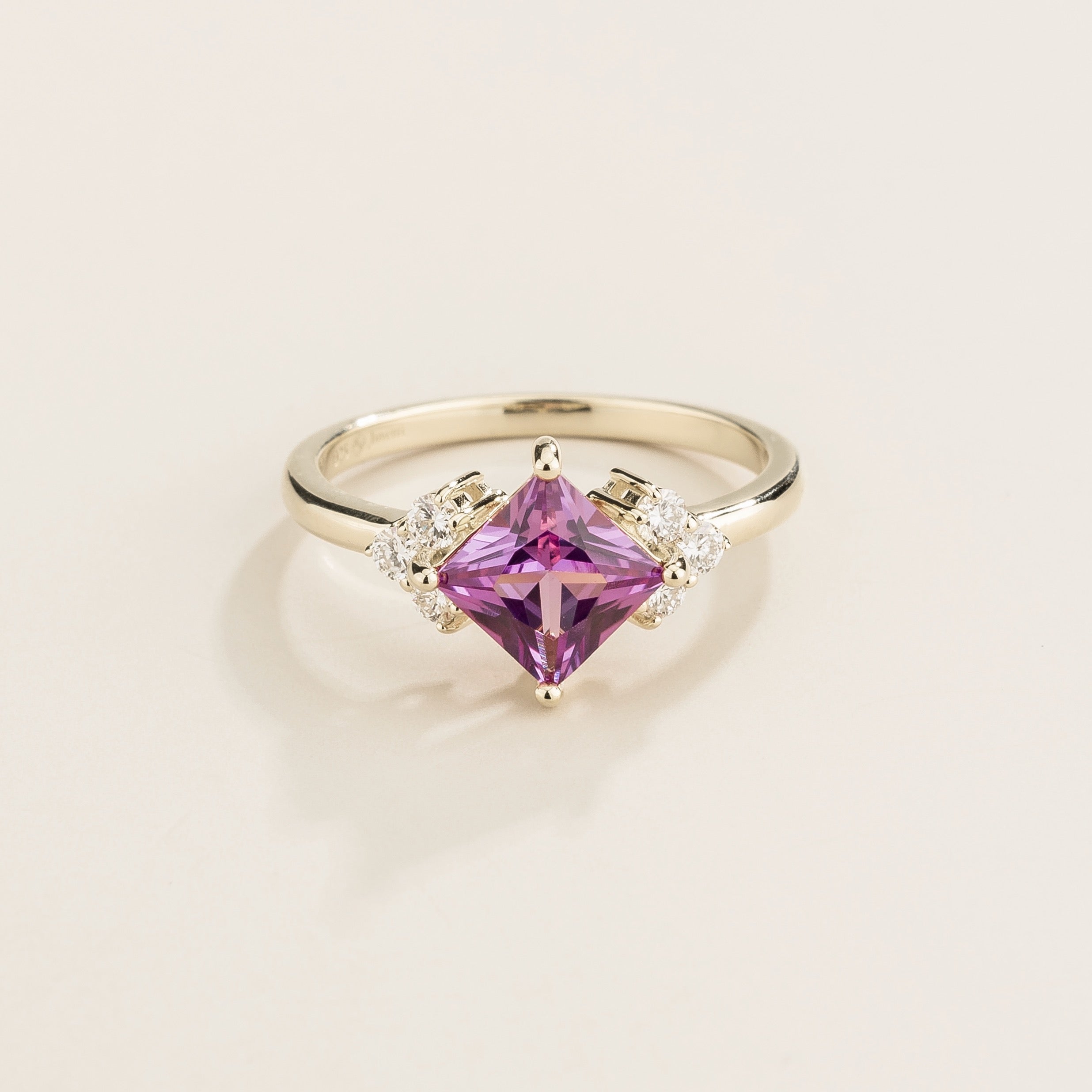 A white gold ring featuring a central princess cut purple sapphire with smaller white diamonds on either side.