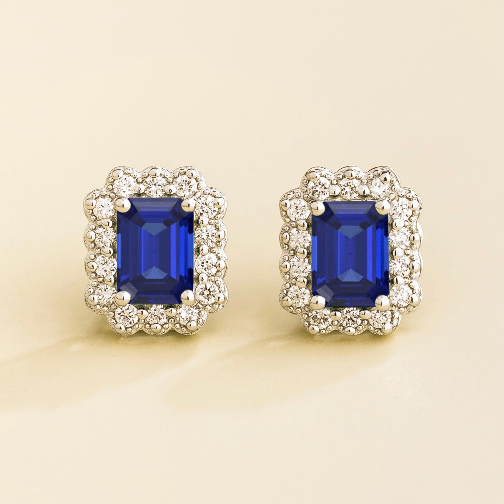 A pair of white gold earrings featuring royal blue sapphires in emerald cuts, surrounded by halo diamonds.