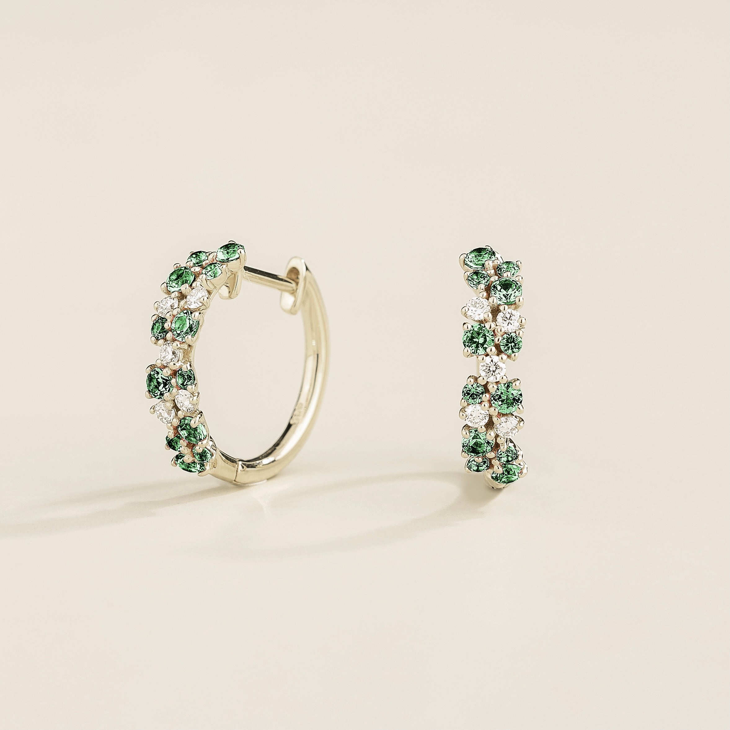 A pair of white gold huggie earrings set with cluster of round emerald and diamond accents, displayed against a neutral background.