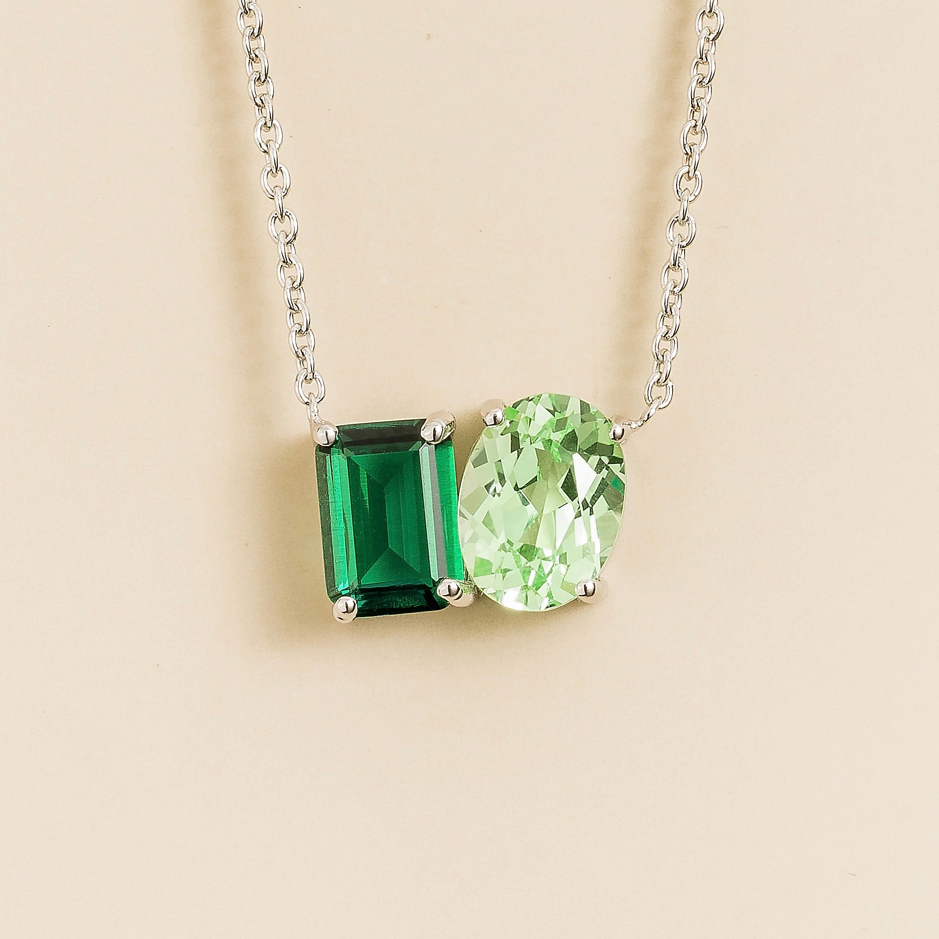 White gold Toi et Moi necklace with an emerald cut green emerald and an oval green sapphire pendant on a light background.