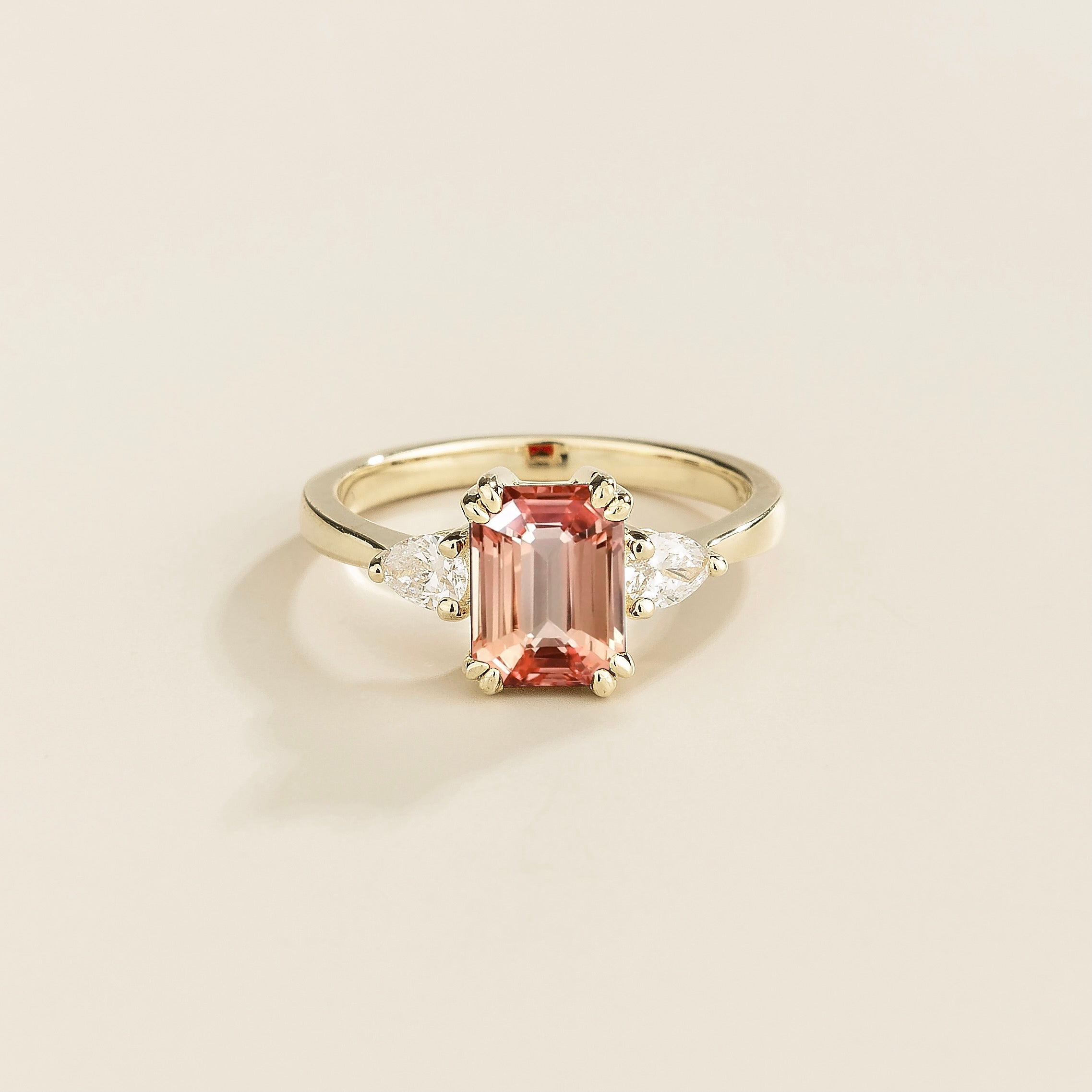 A gold ring with a padparadscha sapphire center stone and pear shape diamonds set in white gold, emerald/octagon cut for the sapphire.