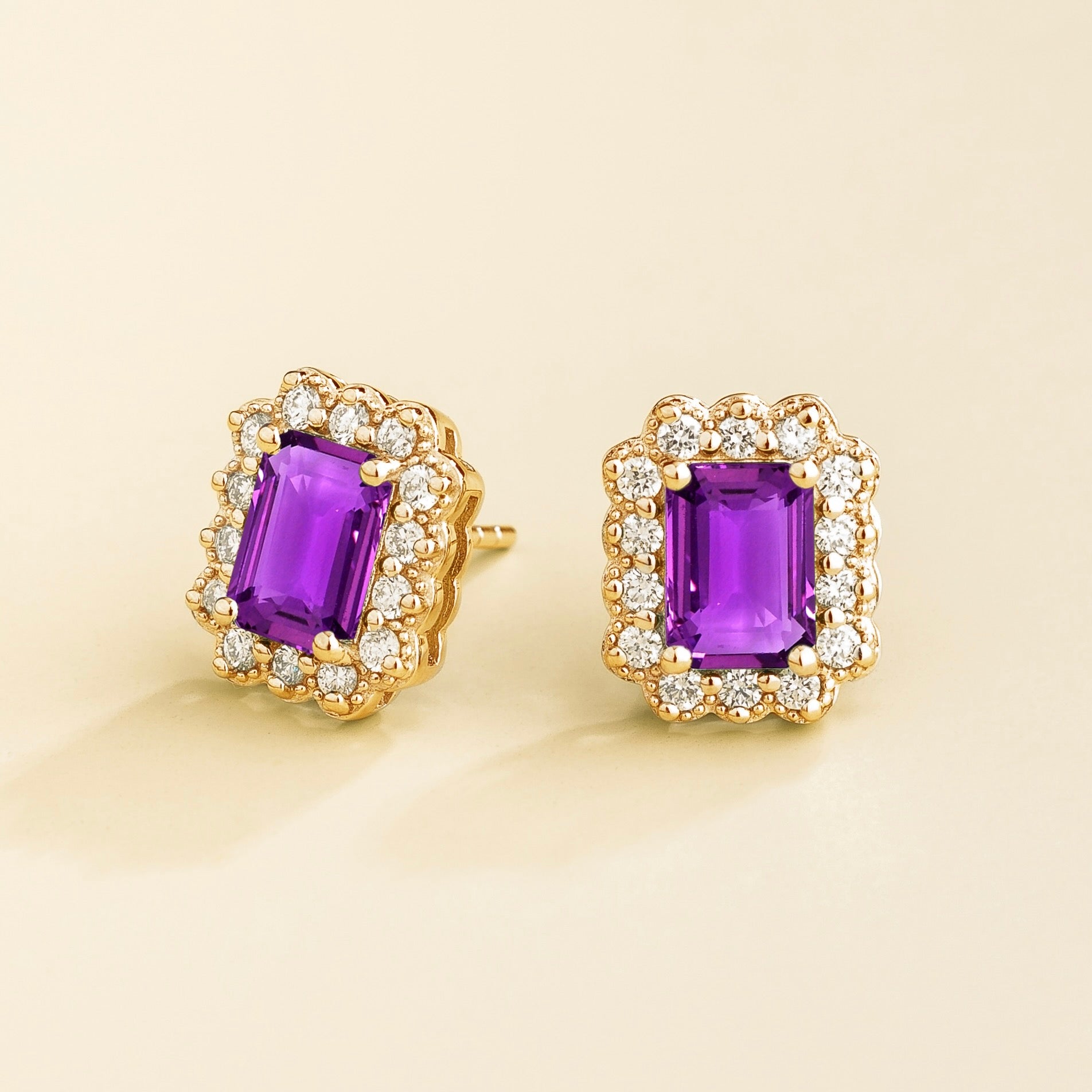 A pair of gold earrings featuring emerald cut purple sapphire centers with halo diamonds around them, displayed against a light background.