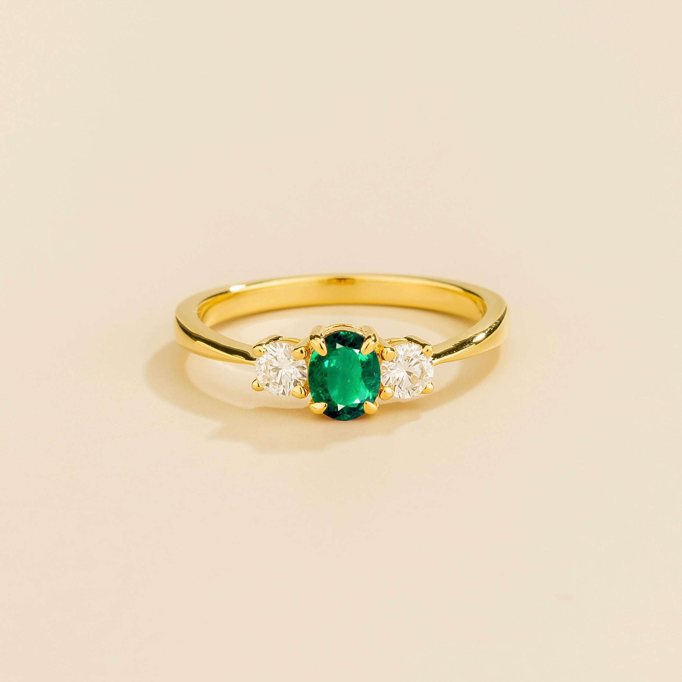 A 3 gemstone gold ring featuring a central emerald flanked by two diamonds on a yellow metal band, presented against a beige background.