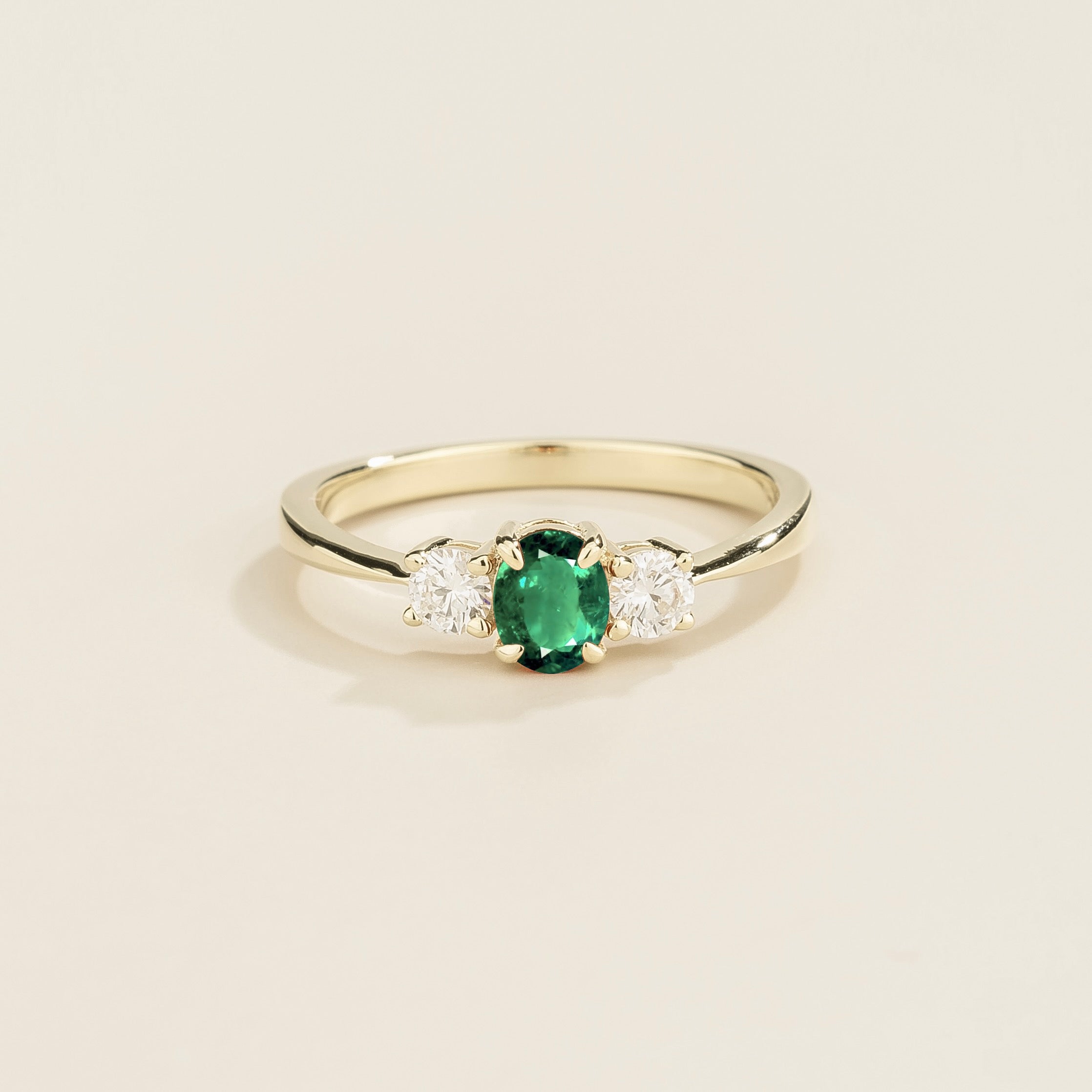 A white 3 gemstone gold ring with an emerald center stone flanked by two smaller diamond stones on either side, set against a light background.