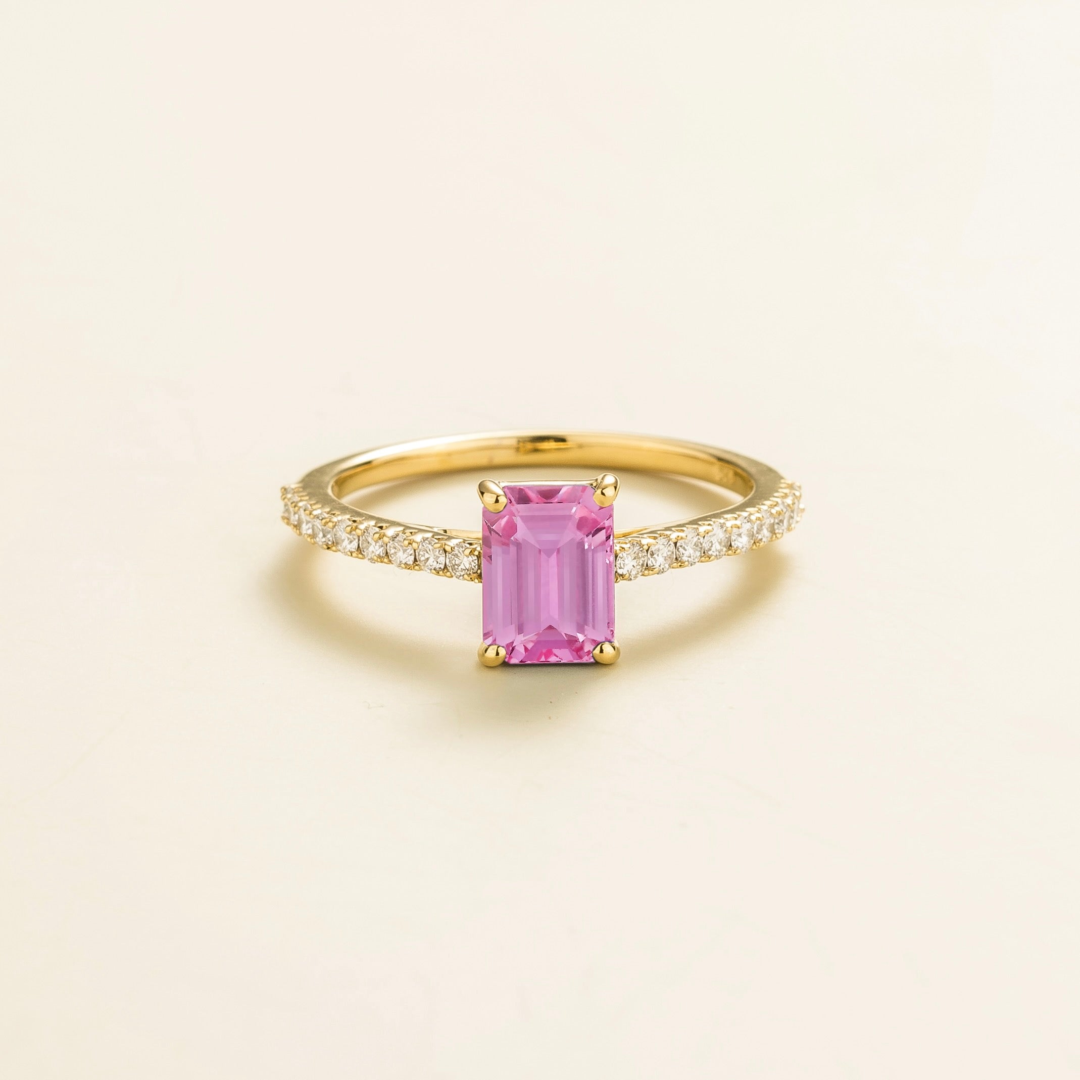 A gold ring with an emerald-cut pink sapphire center stone and smaller diamond accents along the band, displayed against a light background.
