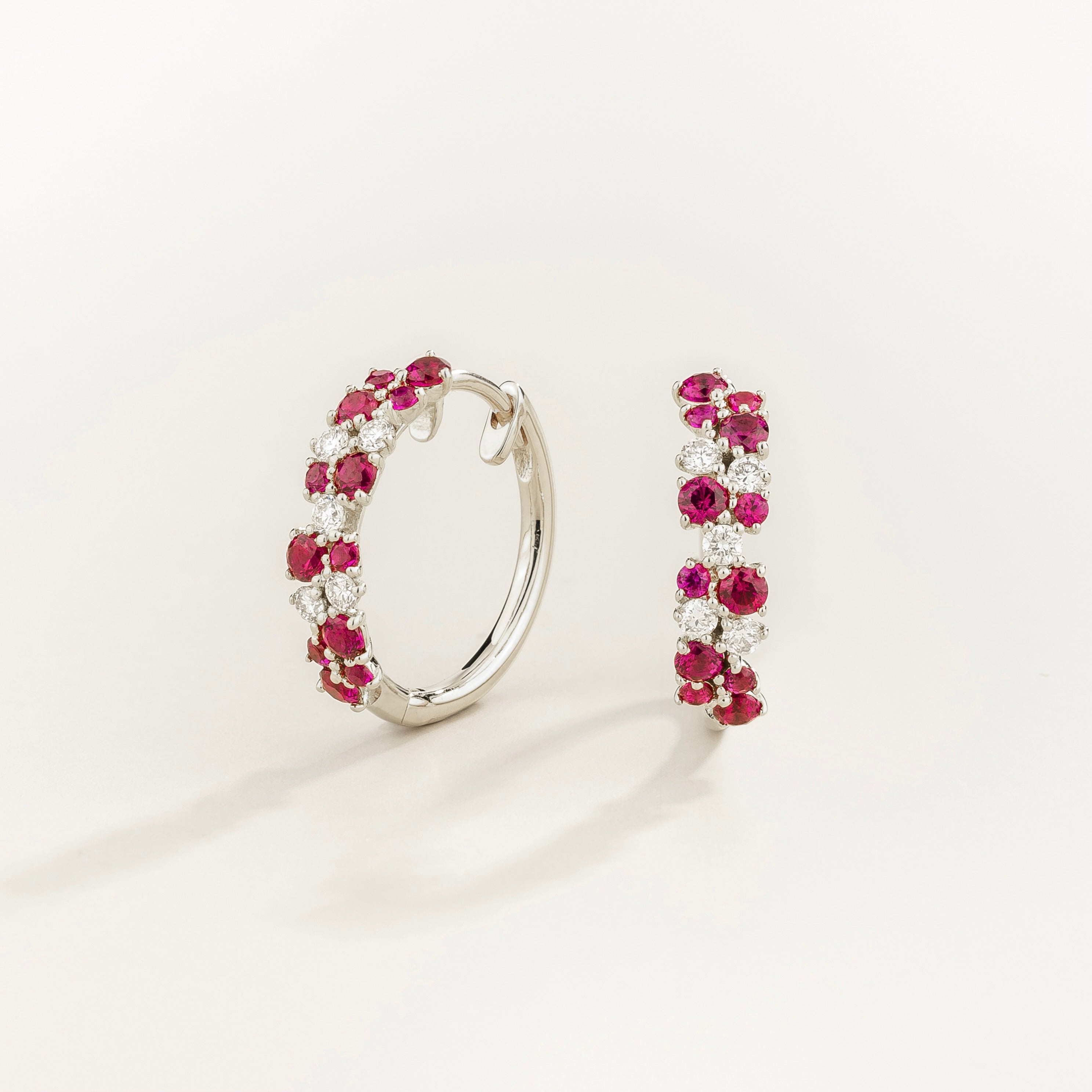 A pair of white gold huggie earrings set with cluster of round ruby and diamond accents, displayed against a neutral background.