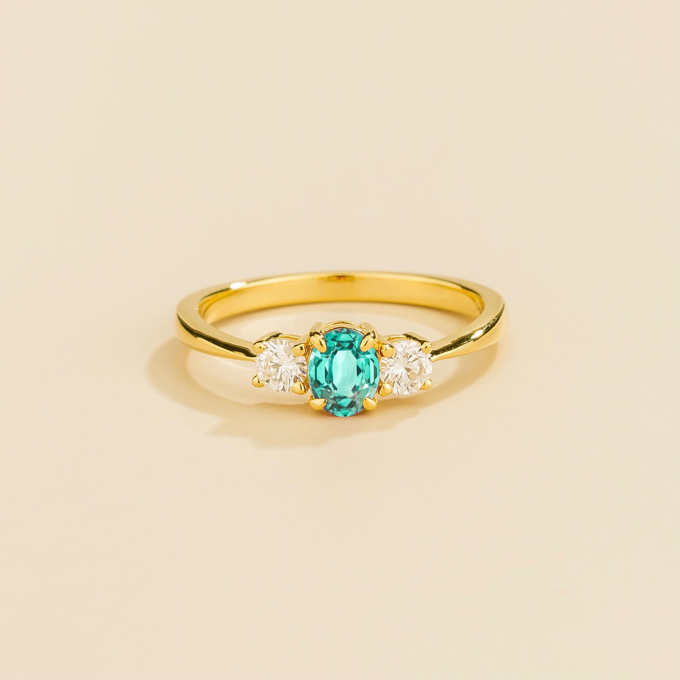 A gold 3 gemstone ring featuring a central Paraiba sapphire with flanking diamonds, displayed against a beige background.