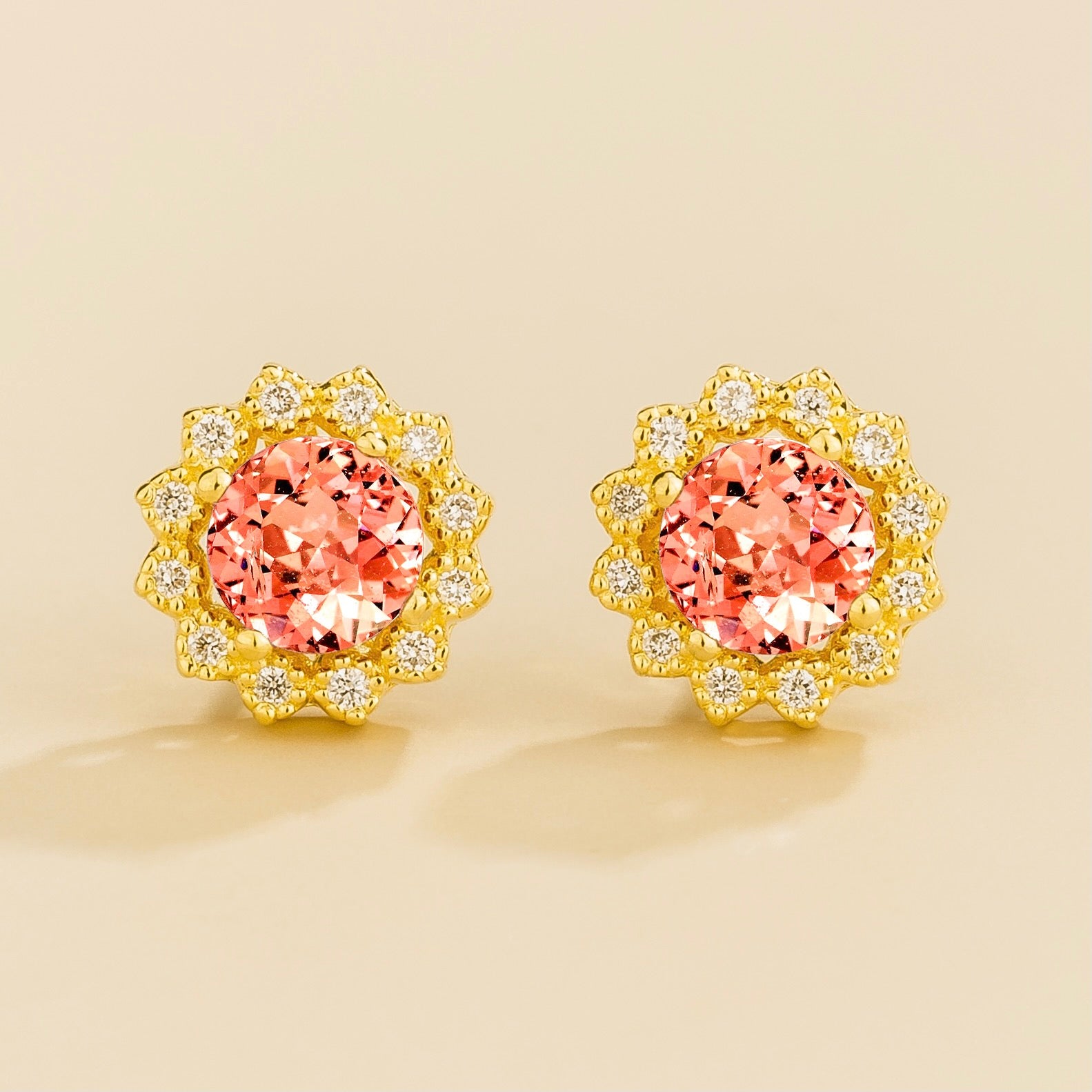 A pair of gold earrings featuring a round cut central padparadscha sapphire surrounded by smaller round diamonds halo. London jewellery store