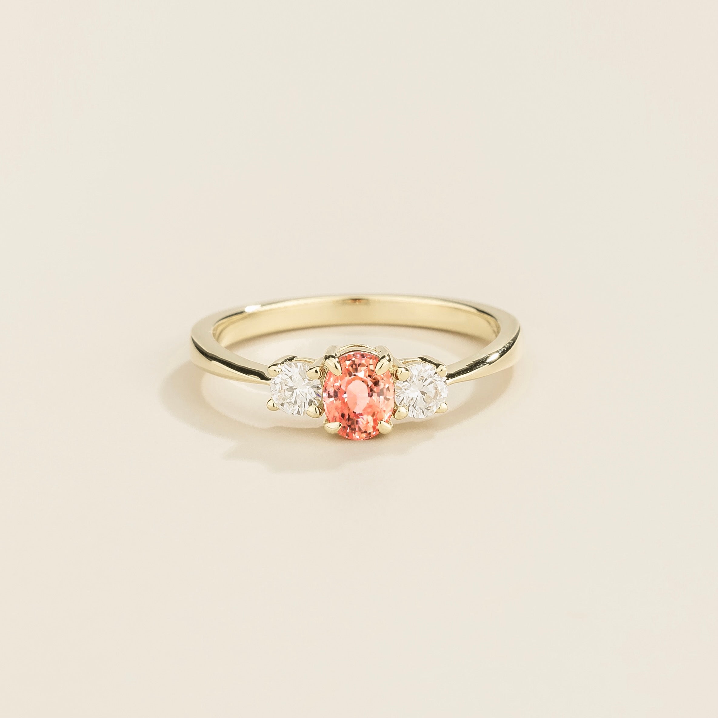 A white gold ring featuring a central Padparadscha sapphire flanked by two diamonds on either side, set against a neutral background.