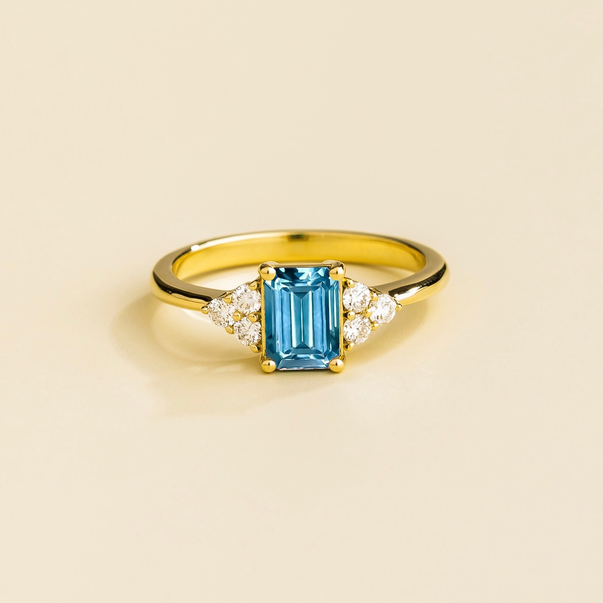 A gold ring with an emerald-cut Aquamarine centerpiece and smaller diamond accents on the sides, displayed against a light background.