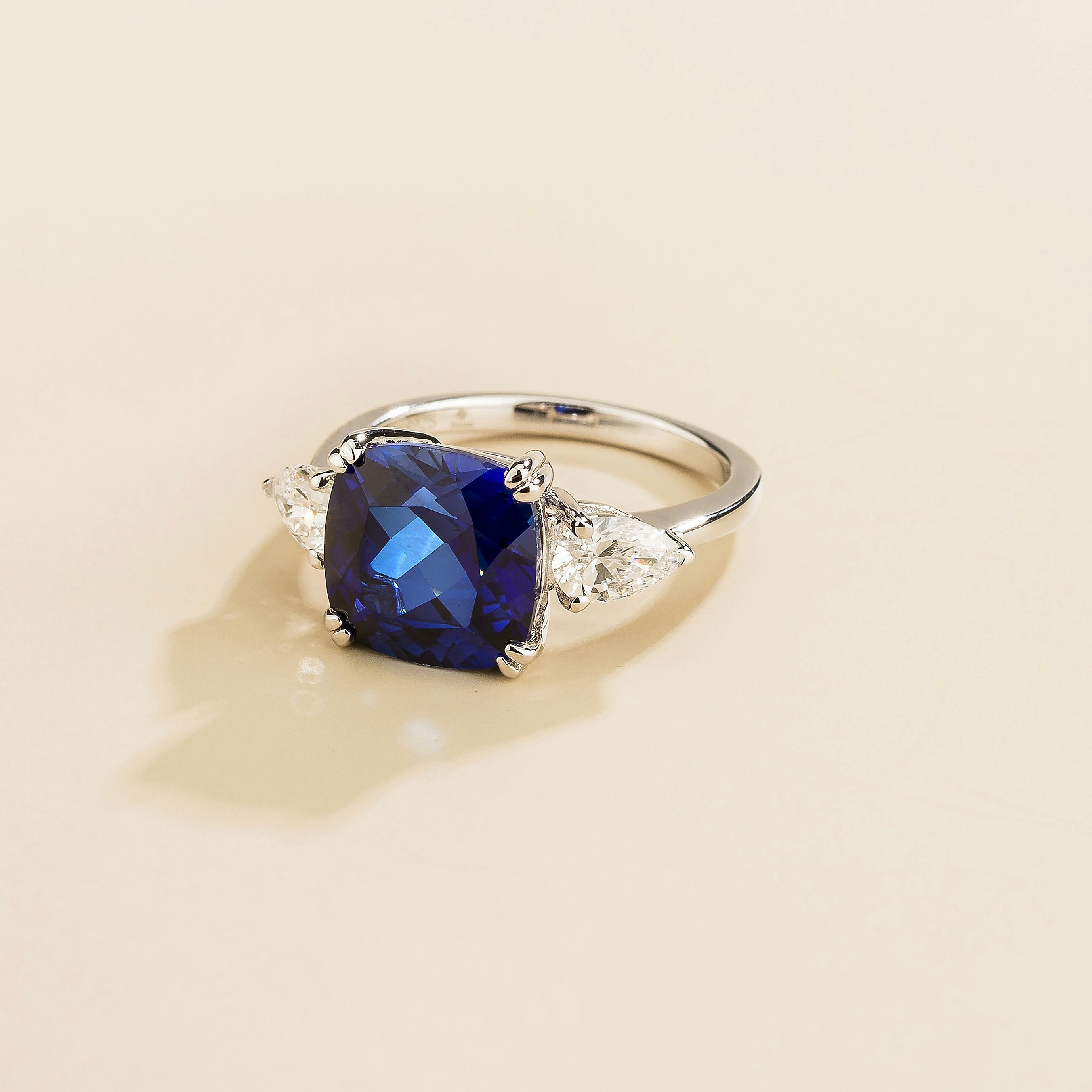 A white gold ring with a large blue cushion-cut sapphire centerpiece and smaller pear-cut diamond accents on the sides.