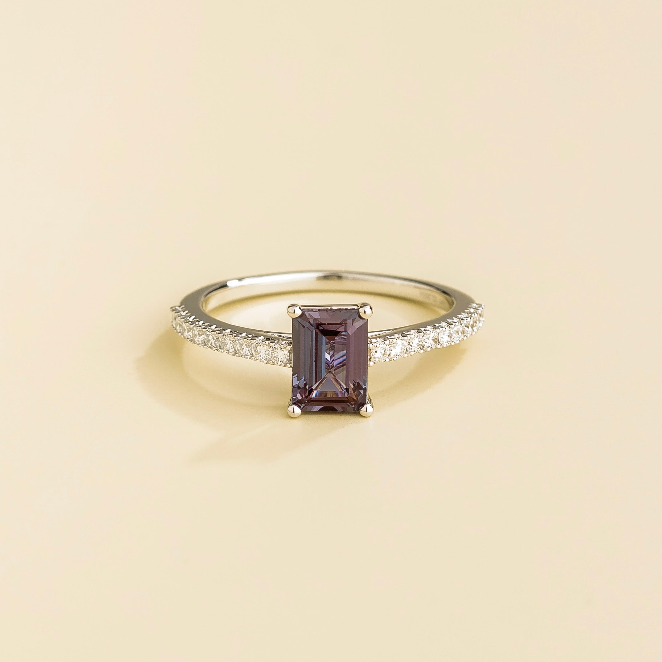 A white gold ring with an emerald-cut alexandrite center stone and pave-set diamonds along the band, displayed against a light background.
