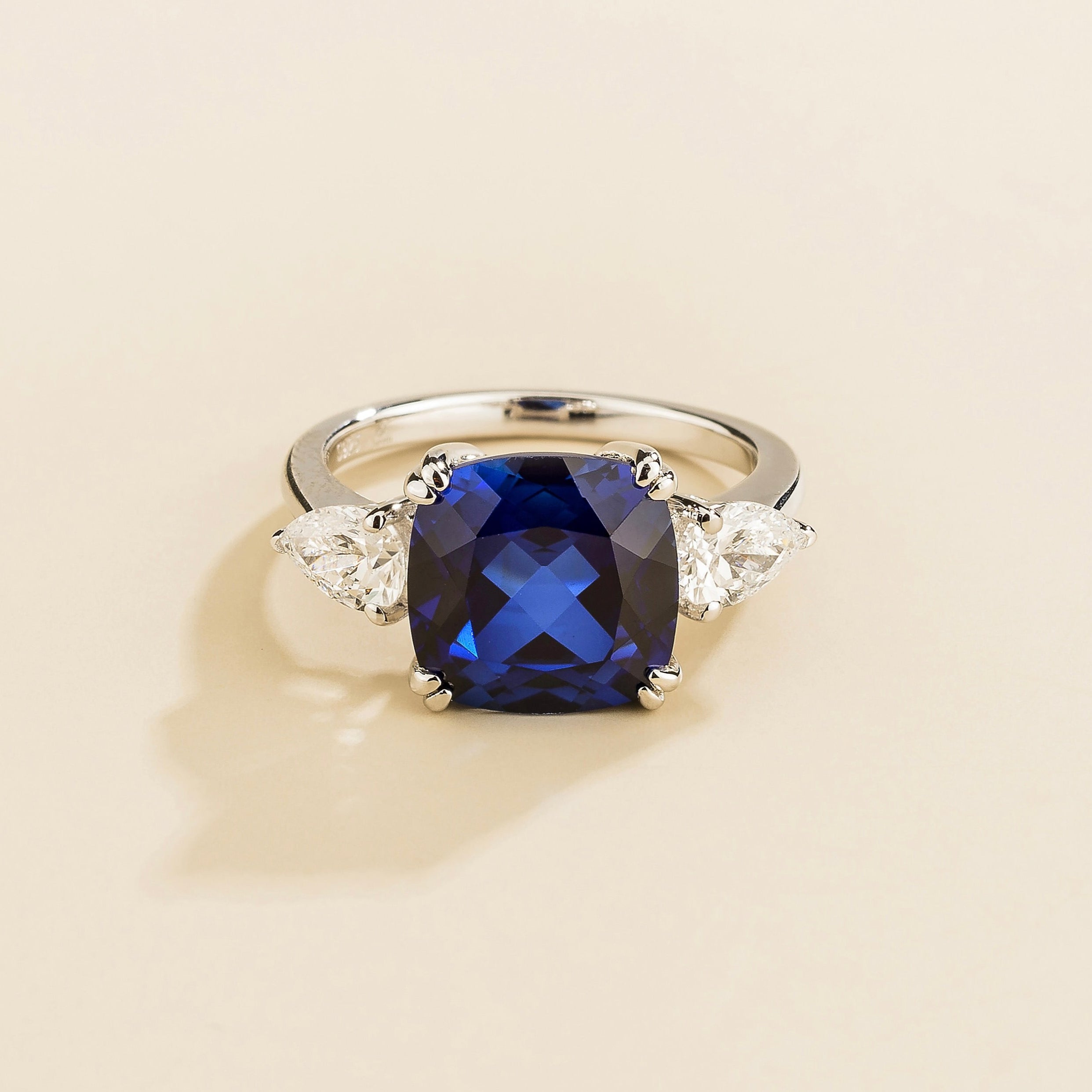 A white gold ring with a royal blue cushion-cut sapphire centerpiece and smaller pear-cut diamond accents on the sides.