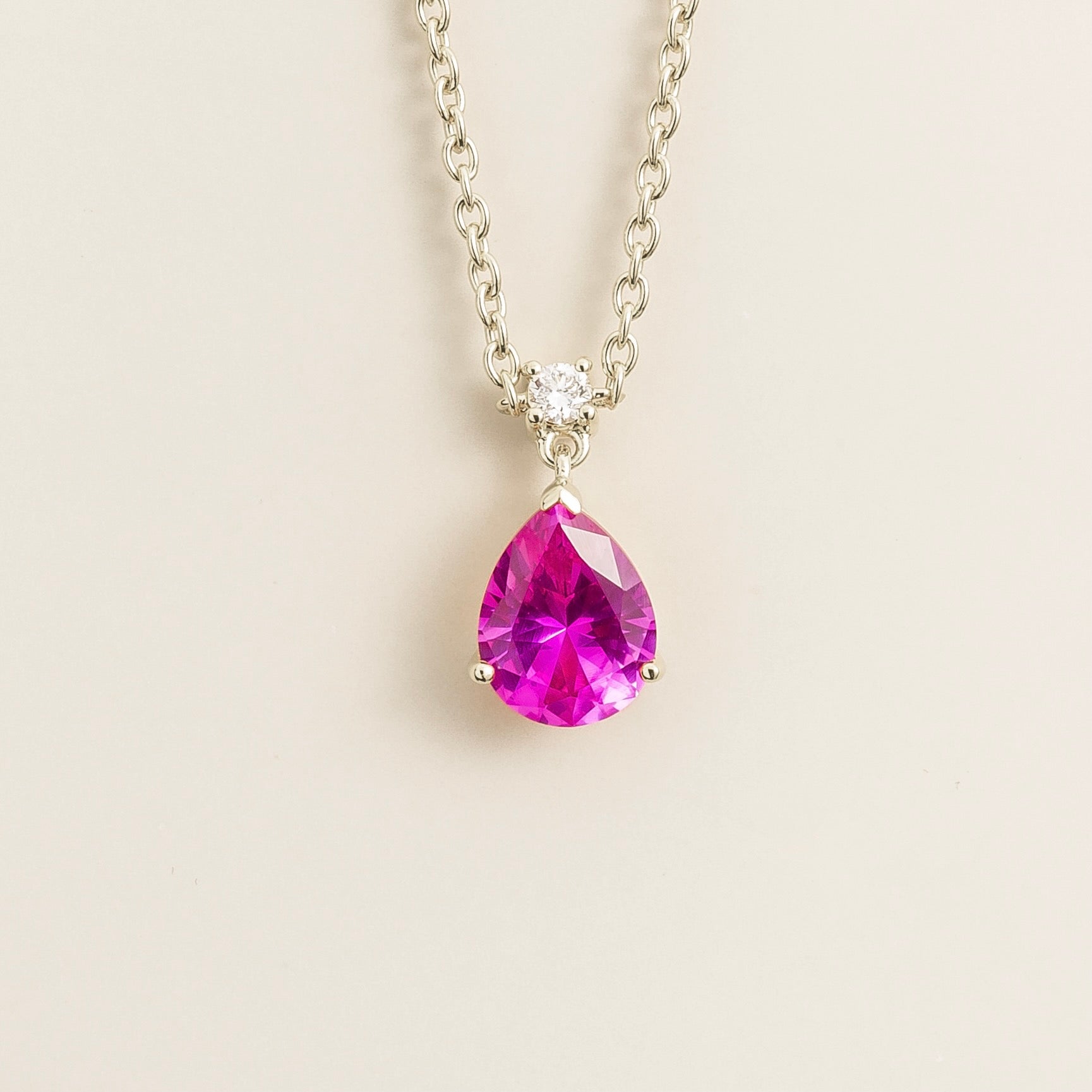 A medium-sized classic pendant necklace in white gold, featuring a pear shape pink sapphire centerpiece and smaller round diamonds, displayed against a neutral background.