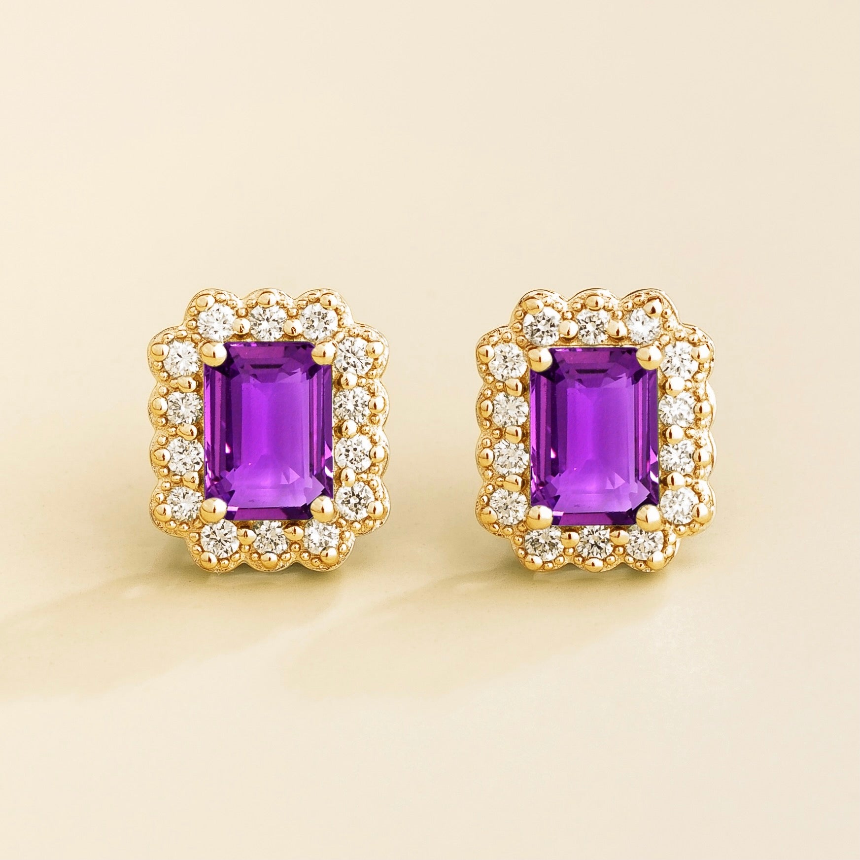 A pair of gold earrings featuring emerald cut purple sapphire centers with halo diamonds around them, displayed against a light background.