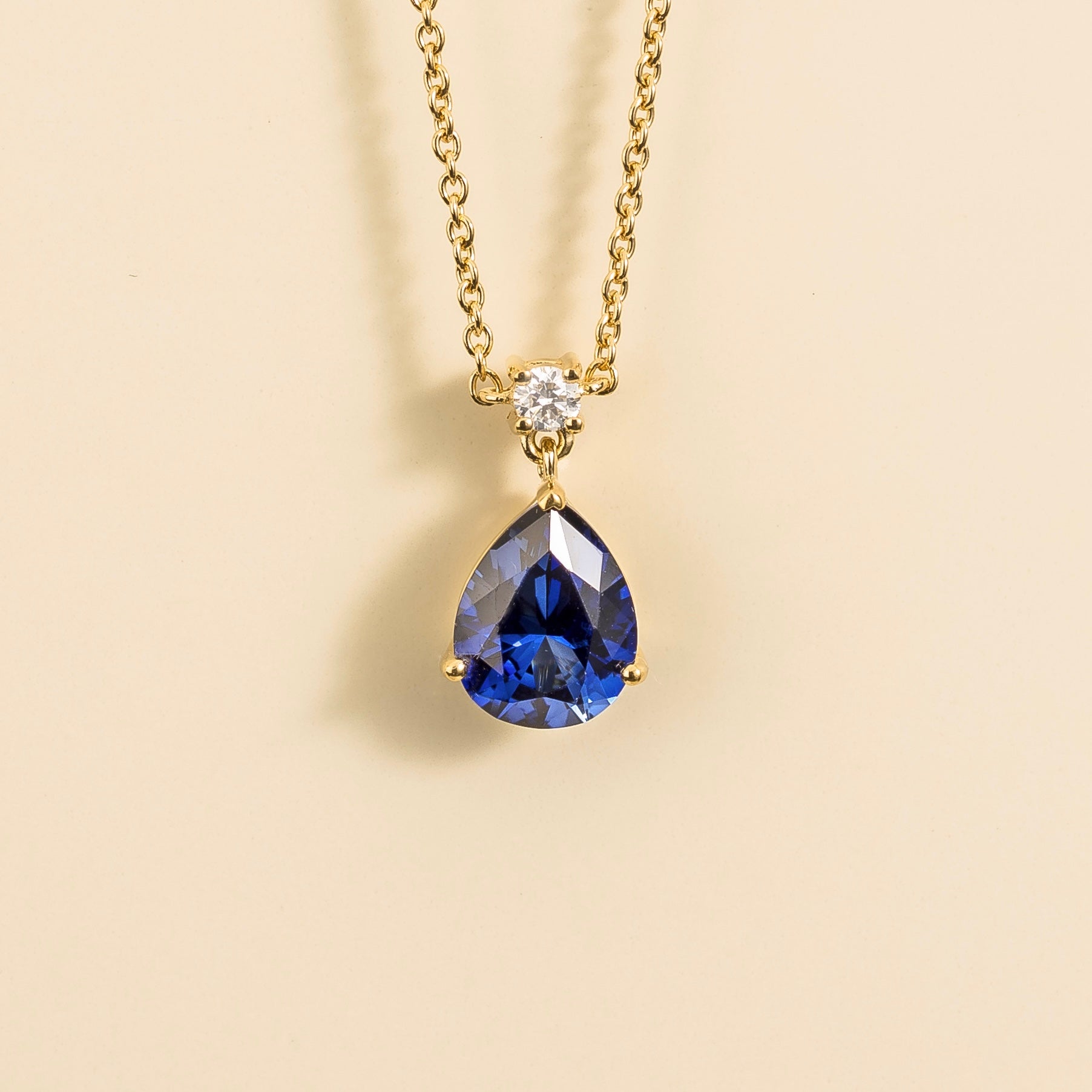 A medium-sized classic pendant necklace in gold, featuring a pear shape blue sapphire centerpiece and smaller round diamonds, displayed against a neutral background.