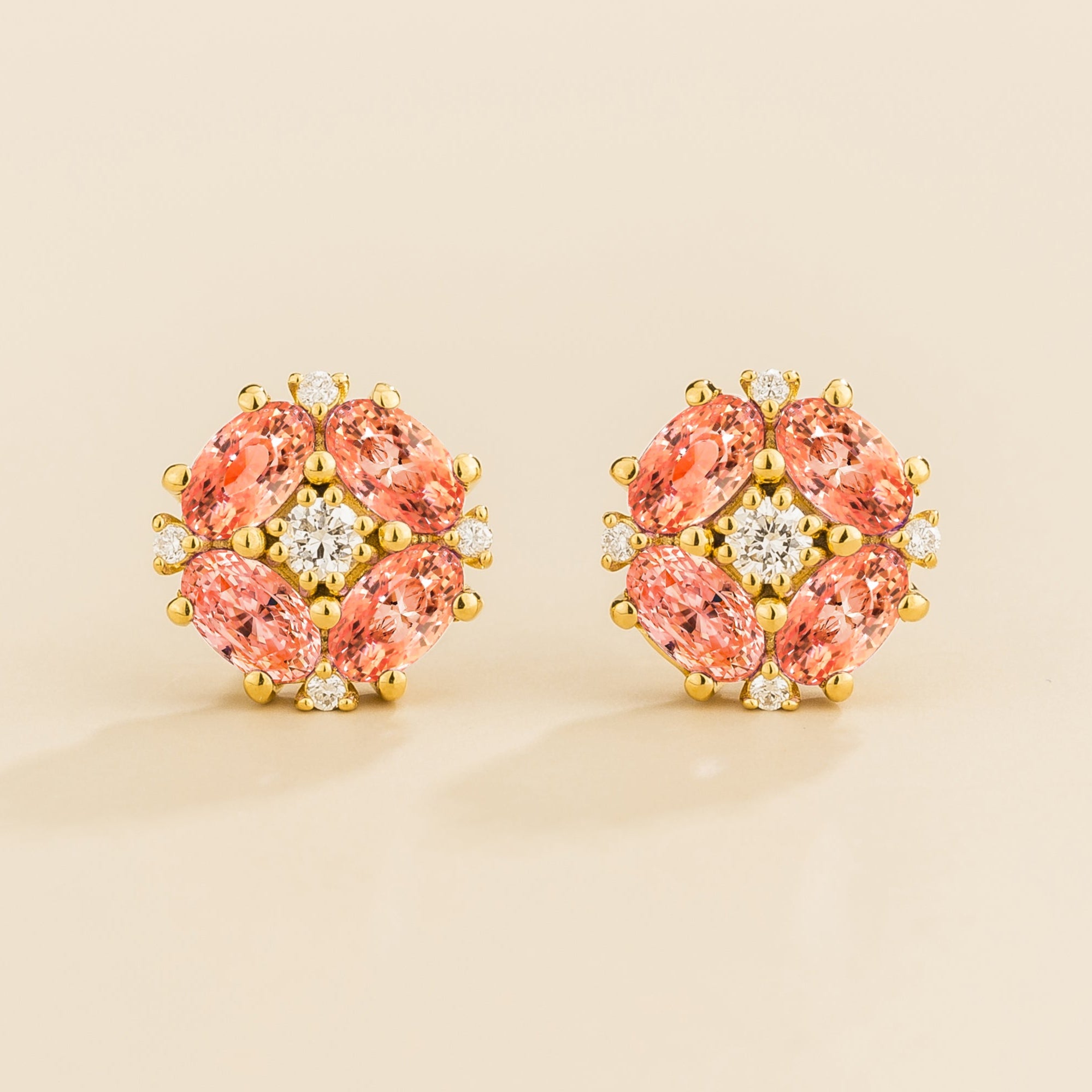 A pair of gold earrings featuring oval padparadscha sapphire and round diamonds arranged in a floral pattern.