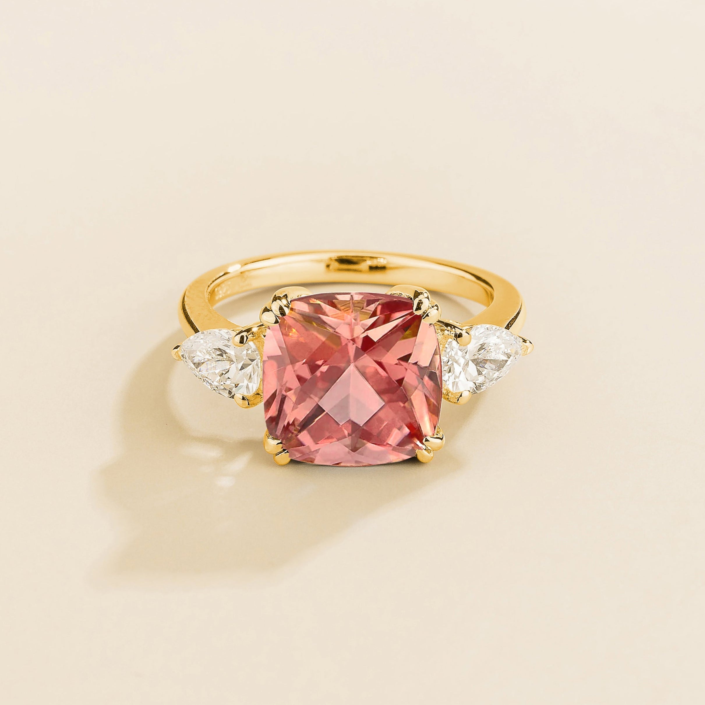 Gold ring with cushion cut padparadscha gemstone and pear side diamonds on a beige background