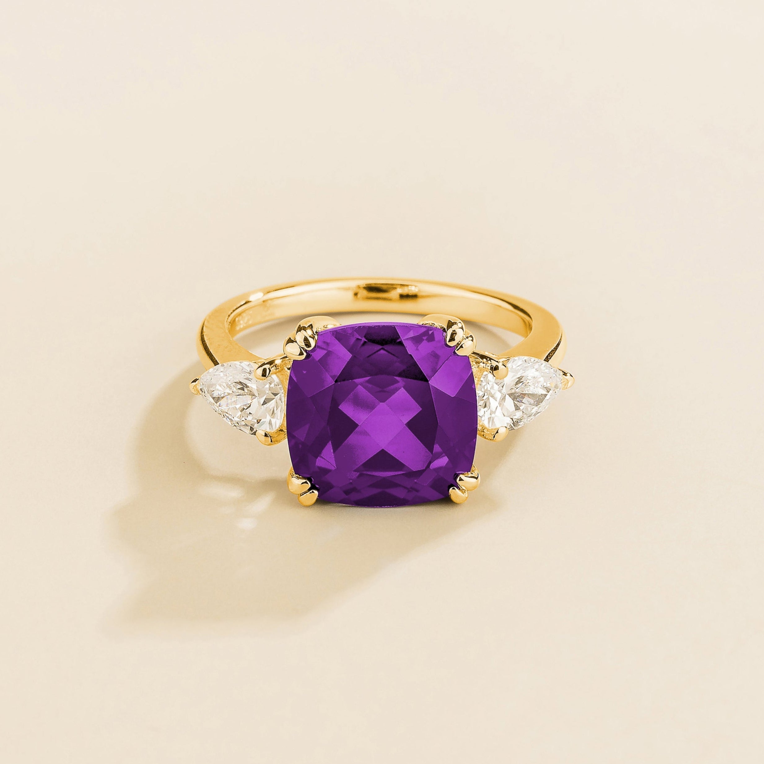 A gold ring with a large purple sapphire cushion cut centerpiece and smaller pear cut diamond accents on the sides.