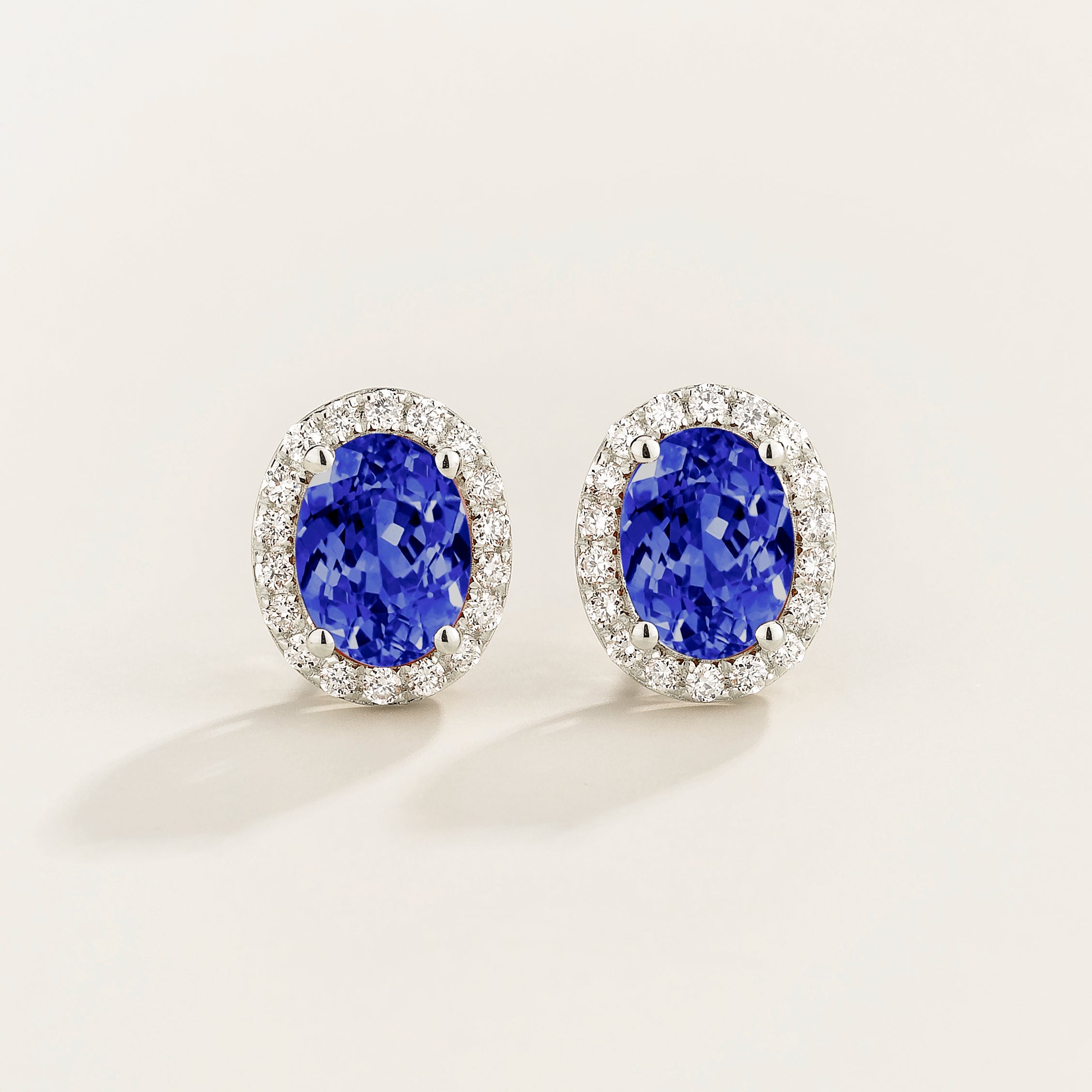Pair of oval blue sapphire gemstone earrings with round diamond halo surrounds on a light background